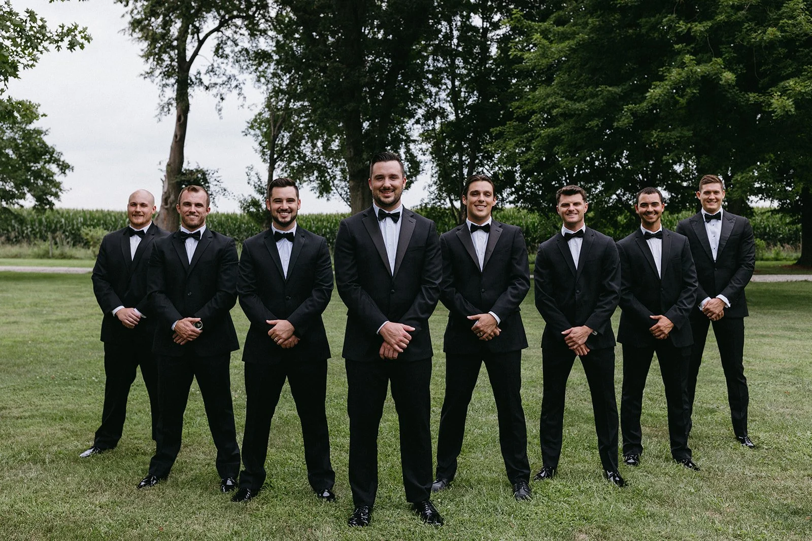 A group of eight men in black tuxedos and bow ties standing outdoors on a grassy field with trees and a cornfield in the background.