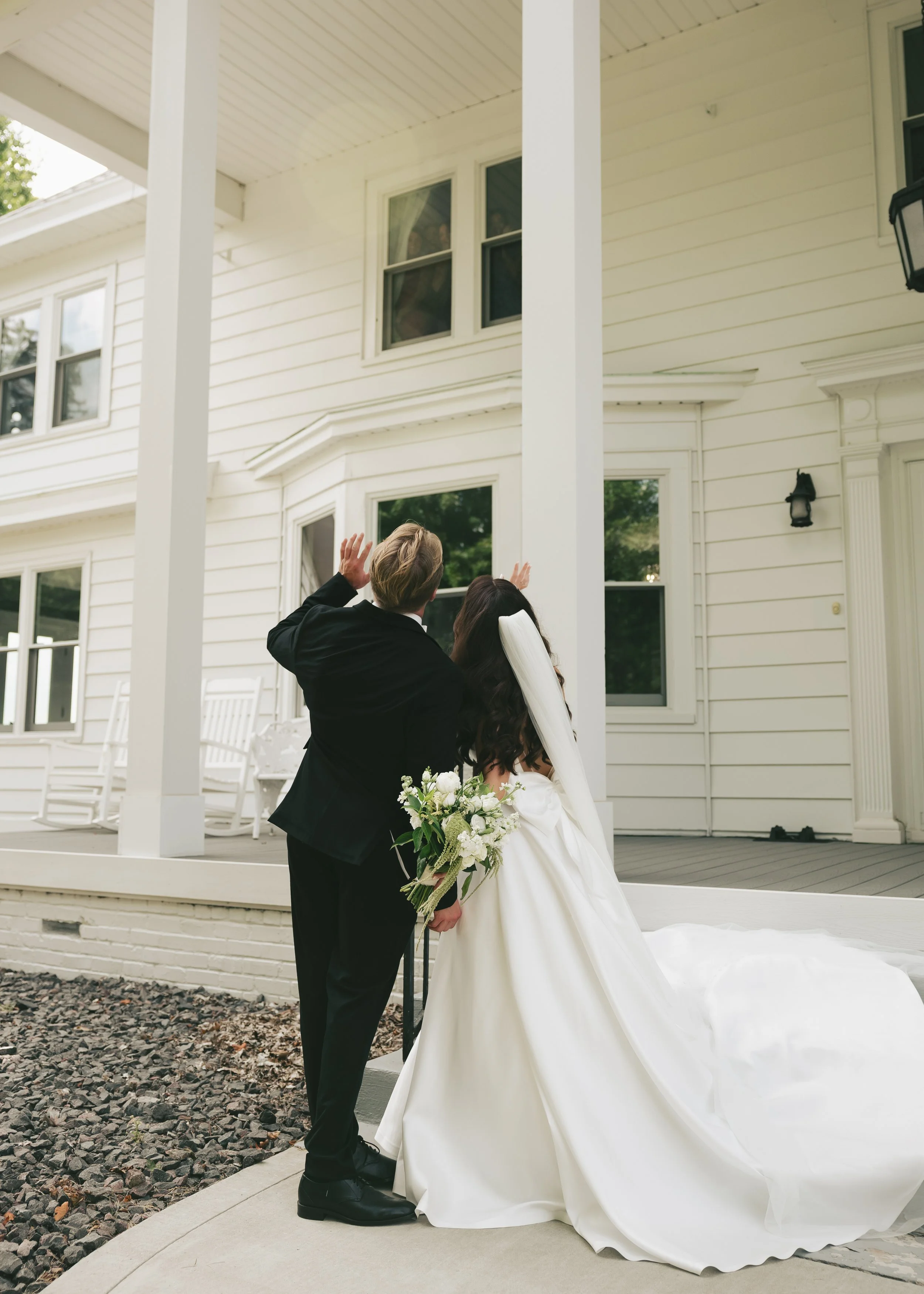 A bride and groom standing outside on a porch, with the bride holding a bouquet of white flowers and wearing a long white wedding dress, while the groom is dressed in a black suit. They are waving to the bridesmaids in the window. 