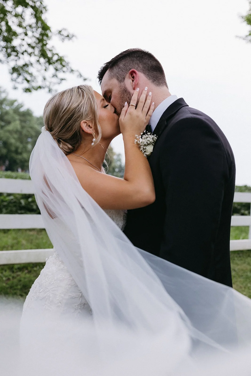 A bride and groom share a kiss outdoors, with the bride in a wedding dress and veil, and the groom in a black suit with a boutonniere. The bride has her hand on the groom's face and a wedding ring visible.
