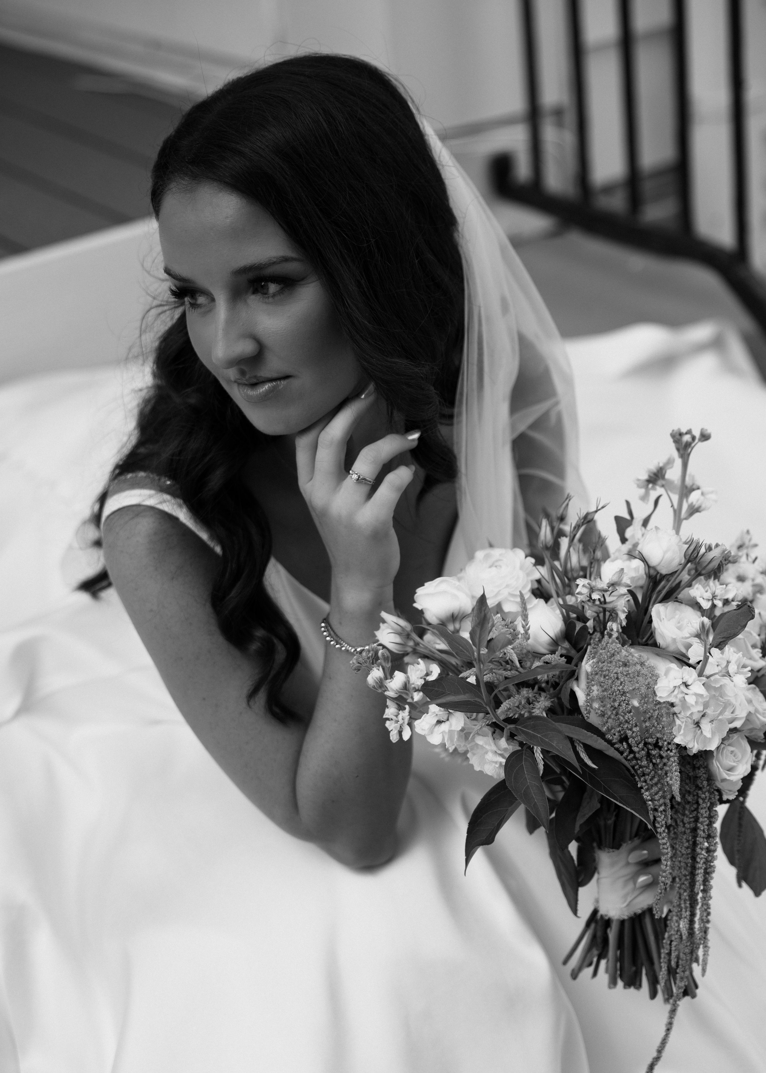 Black and white photo of a woman with long dark hair, wearing a wedding dress, sitting on a porch with a large bouquet of flowers, looking away with a gentle smile.