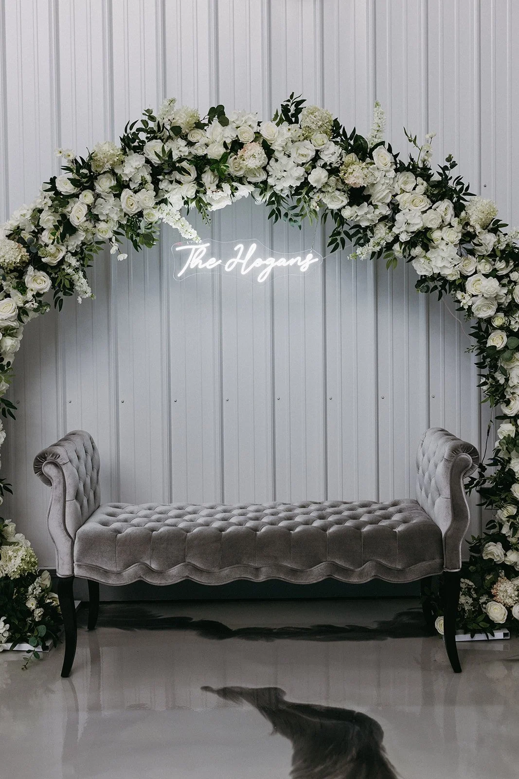 Floral wedding arch with white roses and greenery, illuminated sign that reads 'The Hogans,' a velvet tufted bench, and a white paneled wall.
