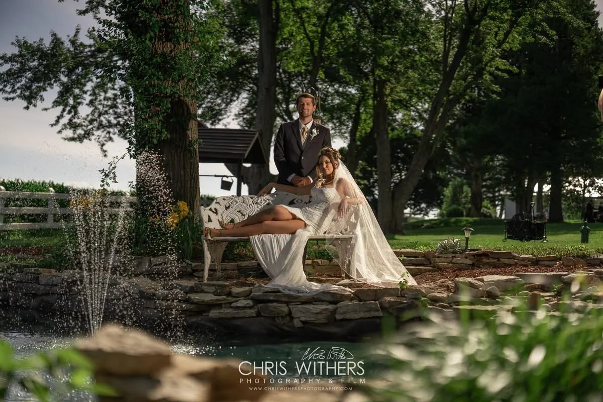 Bride and groom pose outdoors near a pond, with the bride lying on a bench and the groom standing beside her, surrounded by trees and greenery.