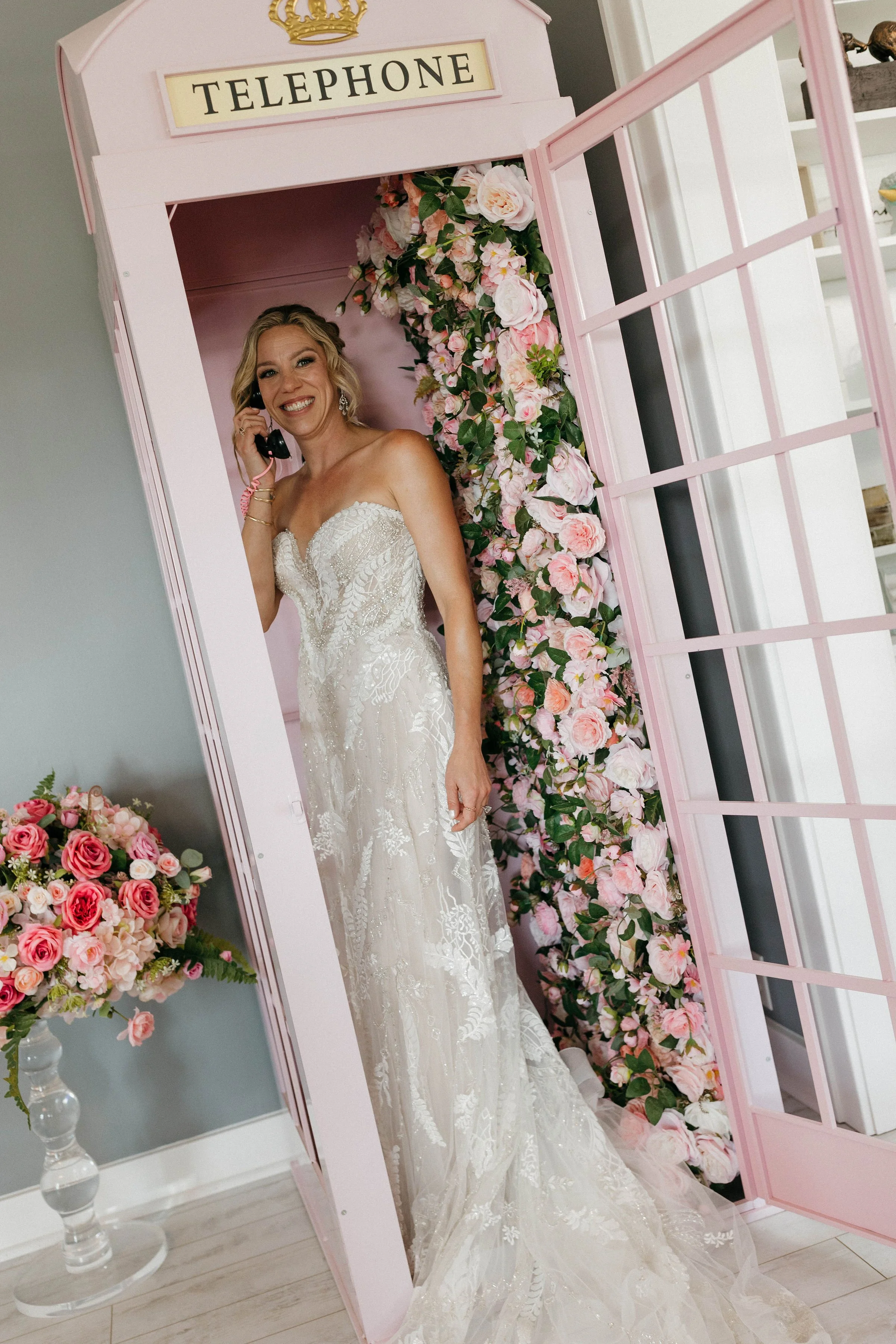 A woman in a wedding dress talking on the phone inside a pink telephone booth decorated with pink and white roses.