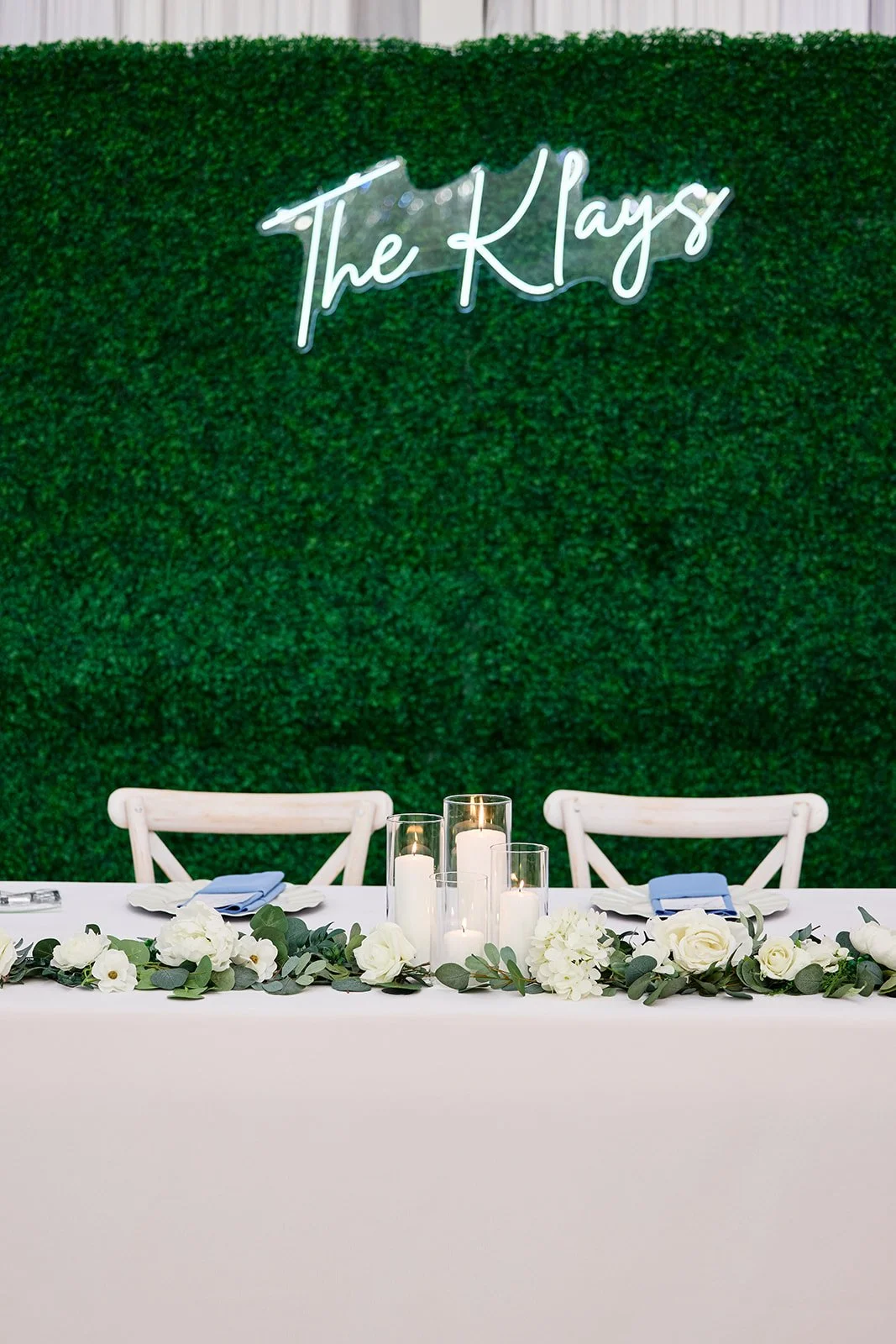 A decorated table with white flowers, candles, and folded cloth napkins in front of a green hedge wall with a neon sign that says 'The Klays'.