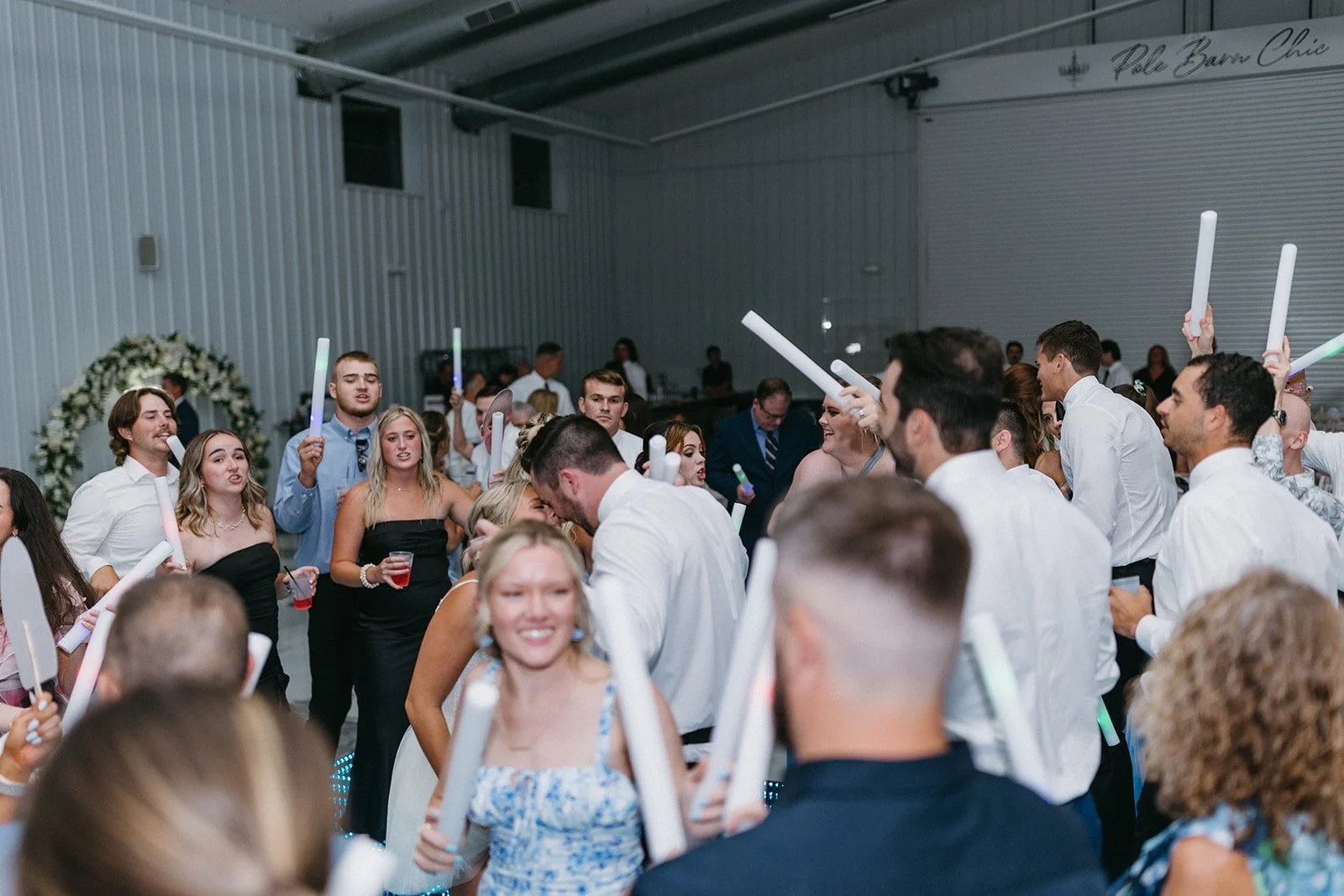 A group of people dancing and celebrating at a wedding reception in a large indoor venue, with some holding glow sticks and drinks, and a floral wedding arch in the background.