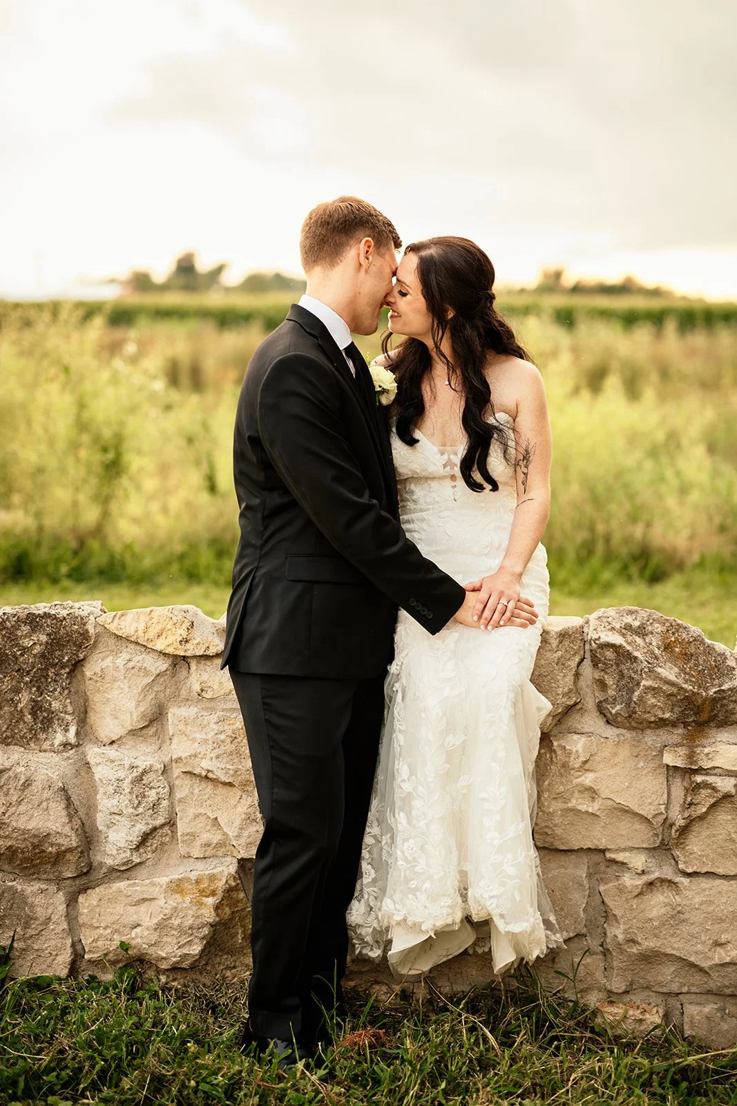 A newlywed couple standing in front of a stone wall in a field, with their foreheads touching and holding hands, dressed in wedding attire, during daytime with overcast sky.