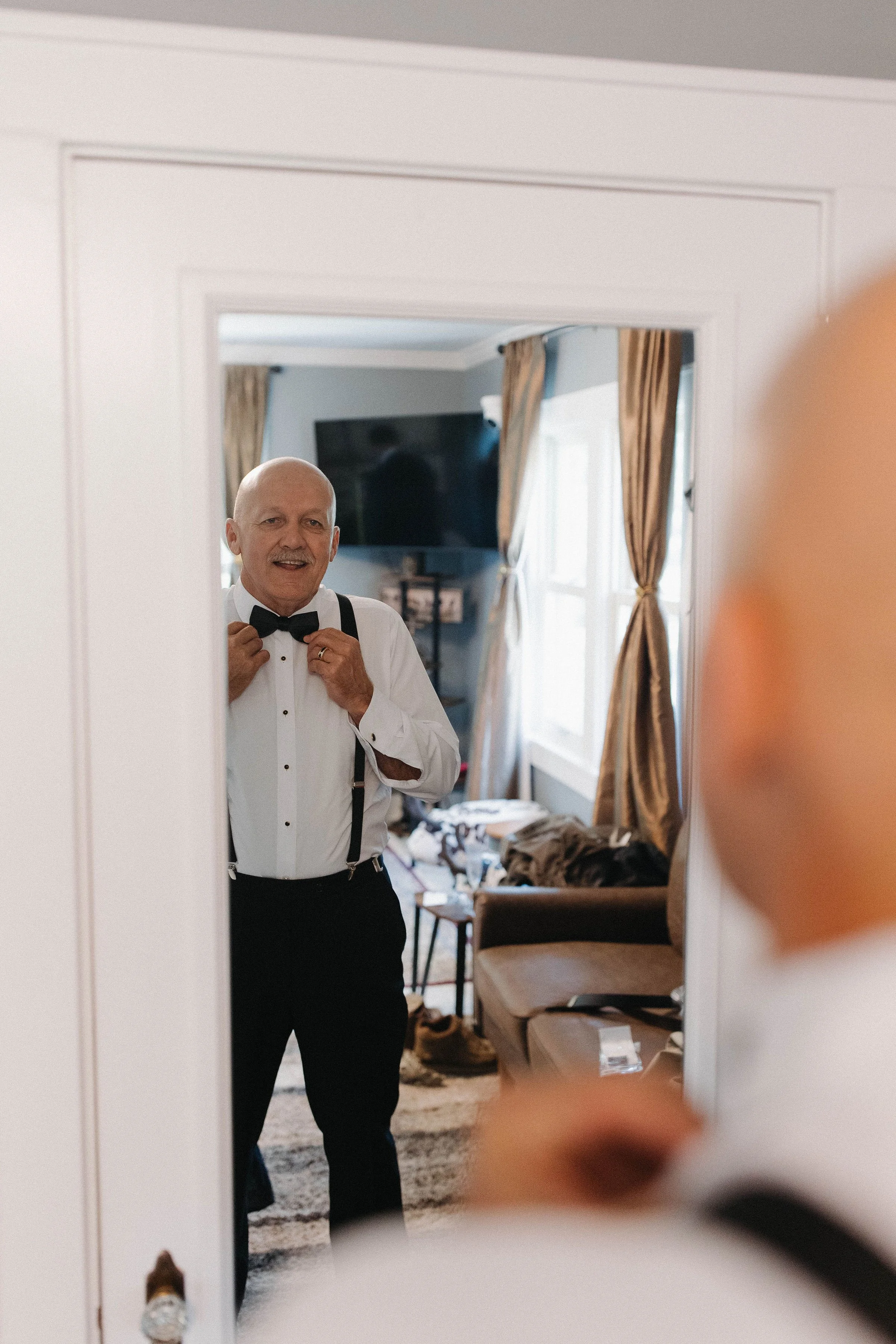 An elderly man getting ready in front of a mirror, adjusting a bow tie, in a warmly lit living room.