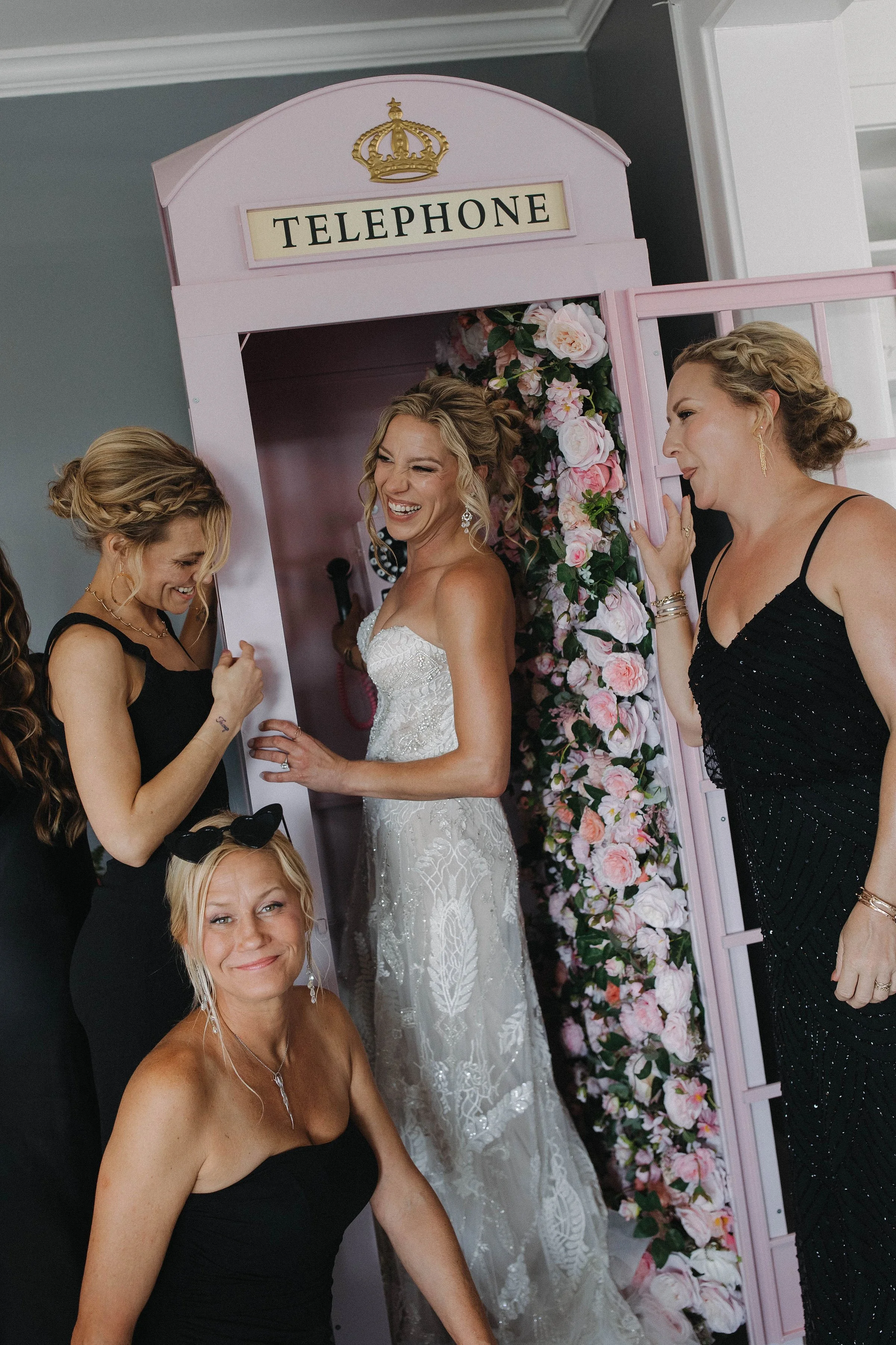 Bride in a white wedding dress surrounded by women in black dress, standing in front of a pink 'Telephone' photo prop decorated with pink and white roses, during a wedding celebration.