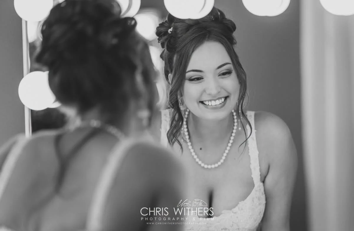 A smiling woman with styled hair, wearing a white dress, pearl necklace, and earrings, looking at her reflection in a mirror.