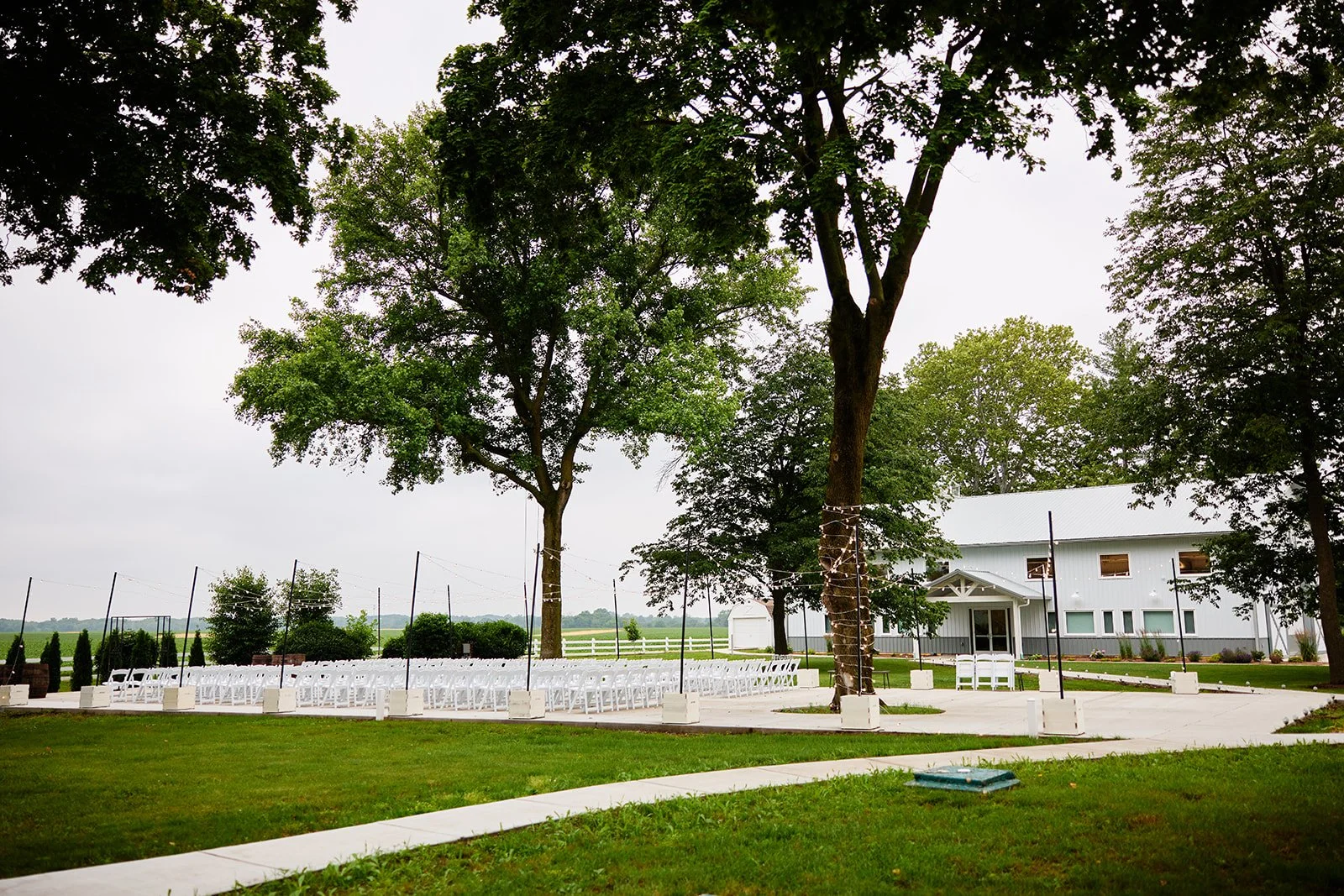 Outdoor wedding ceremony setup with white chairs arranged in rows on a paved area, surrounded by green grass and large trees decorated with string lights, with a white building in the background.