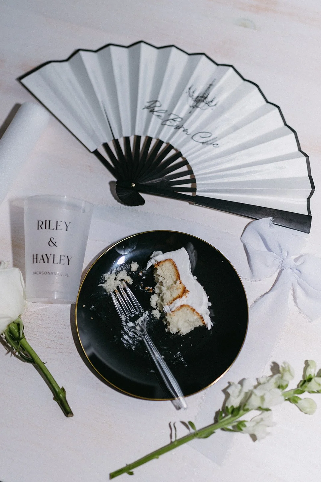 Partially eaten vanilla cake on a black plate with gold rim, silver fork, white napkin, clear cup with text 'Riley & Hayley Jacksonville, IL', paper fan with 'Belle Vue' written on it, white flowers, and a white tablecloth.