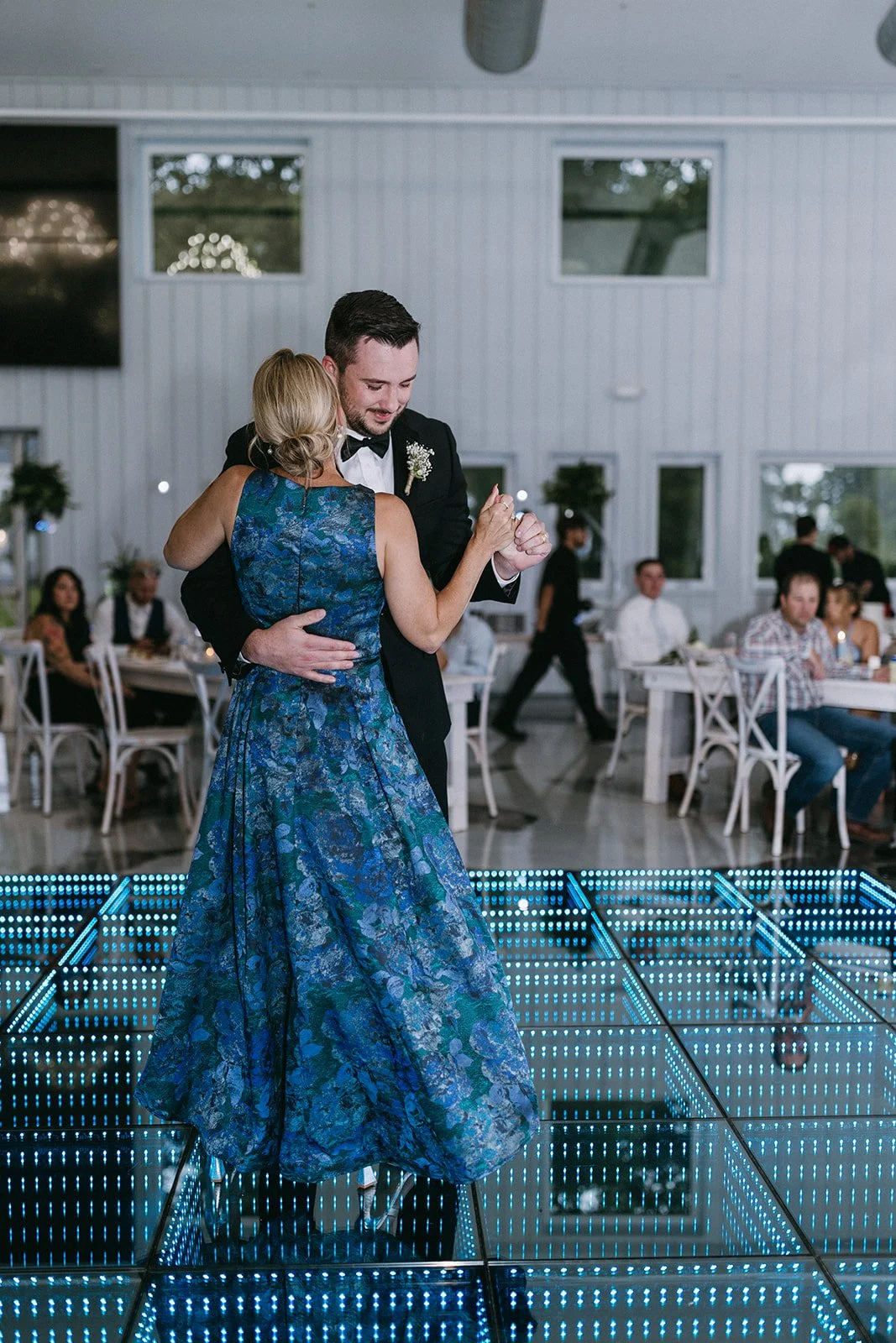 A man and woman dancing at a wedding reception on an illuminated dance floor, with guests seated at tables in the background.
