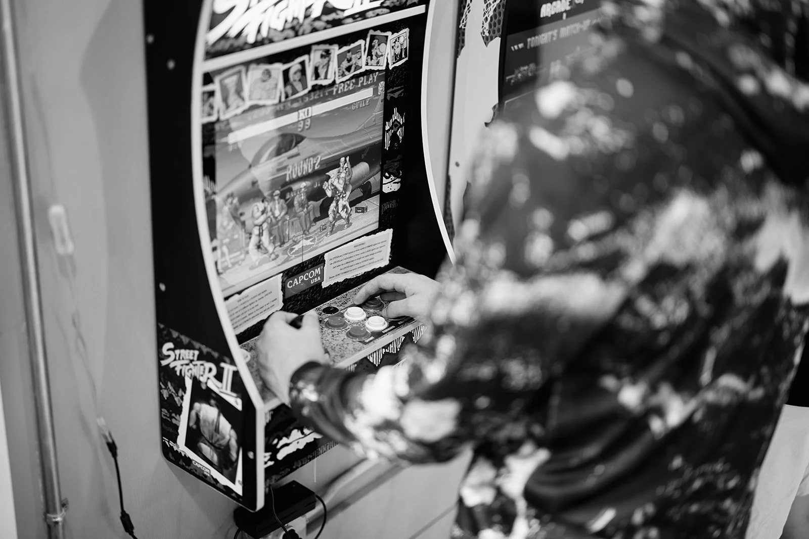 A person playing a Street Fighter arcade game at an arcade. The person's hands are on the control panel, and they are wearing a patterned long-sleeve shirt. The arcade game screen shows a fighting scene from Street Fighter.
