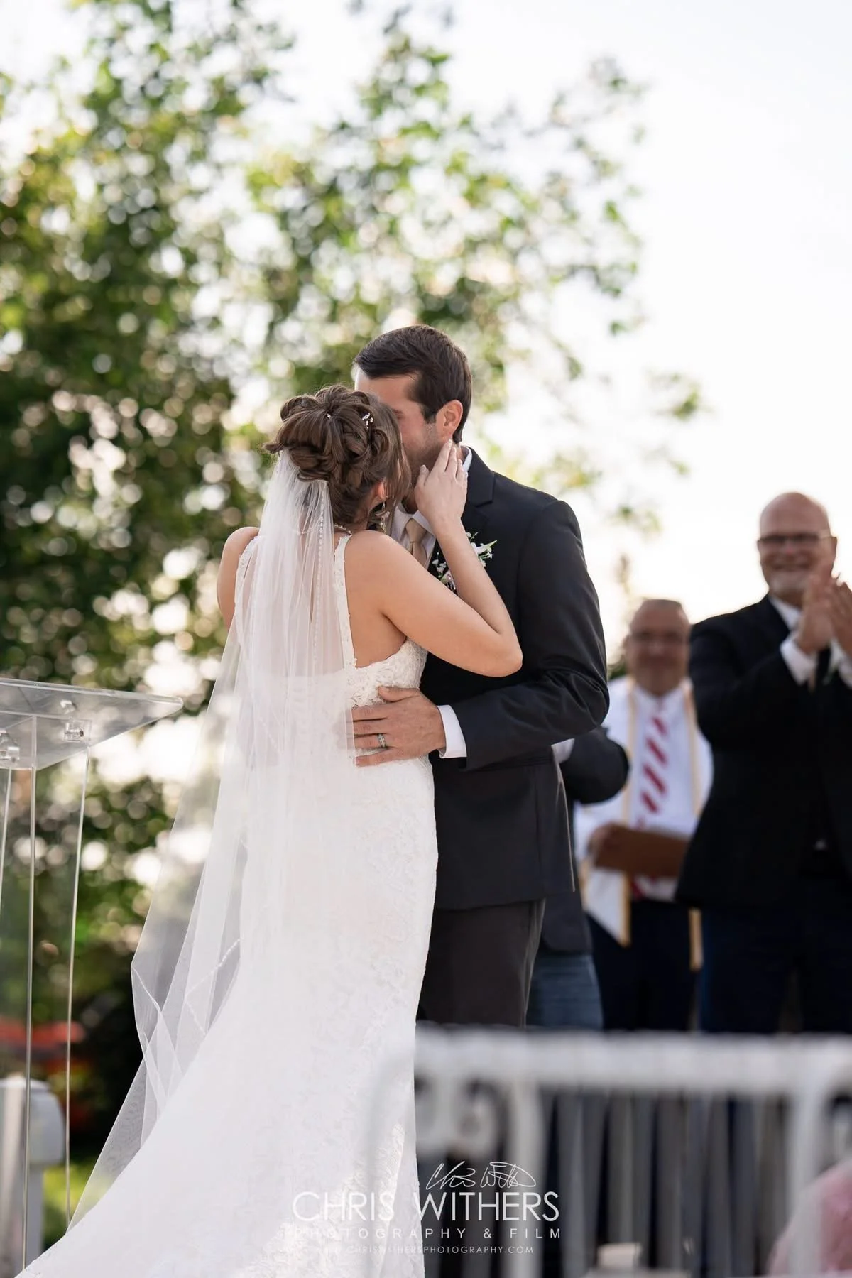 A bride and groom sharing a kiss at an outdoor wedding ceremony, with wedding officiants and guests clapping in the background.
