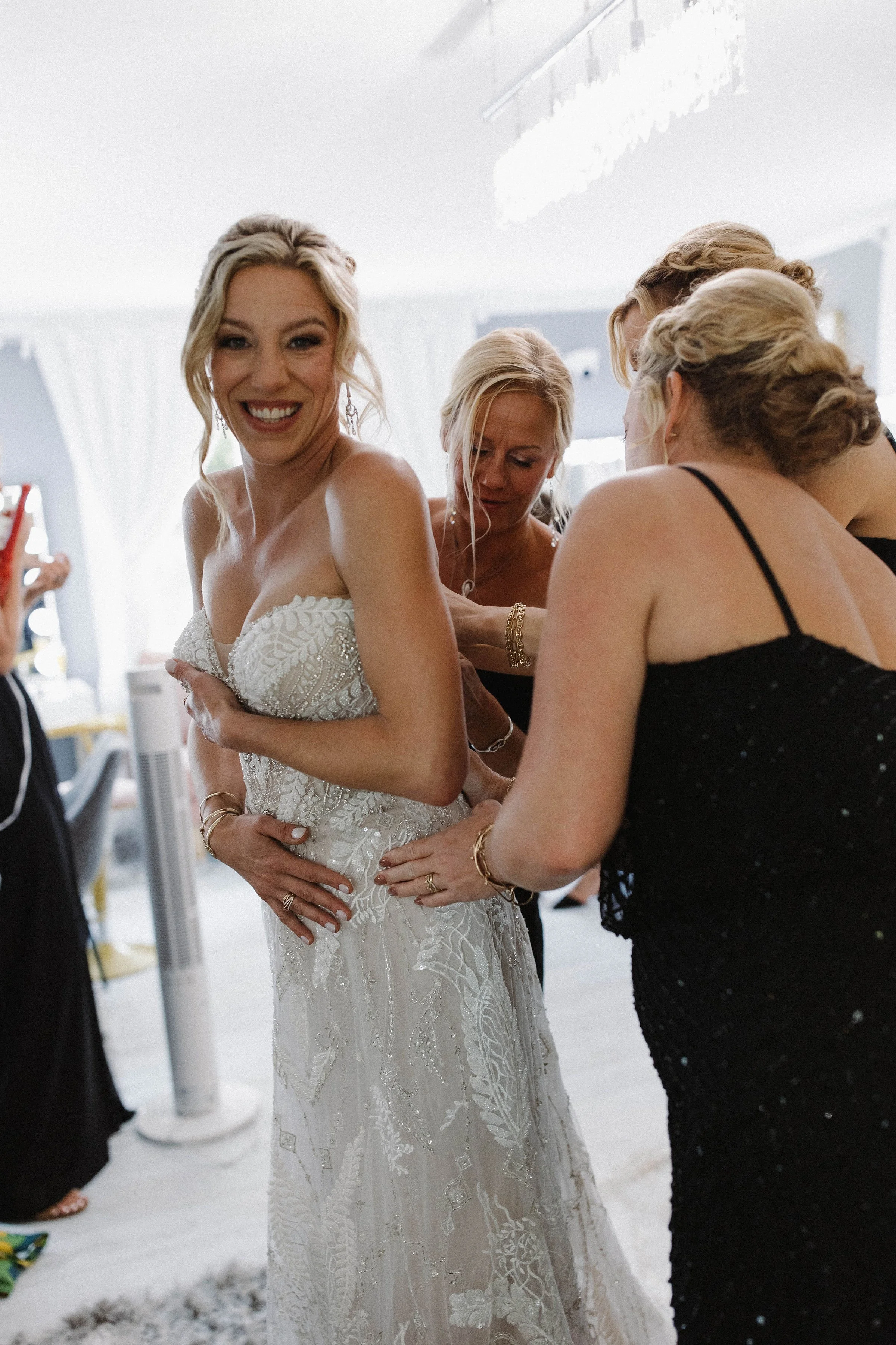 A bride in a white wedding gown getting help from her bridesmaids, with one holding her dress and the others adjusting it, in a bright room.