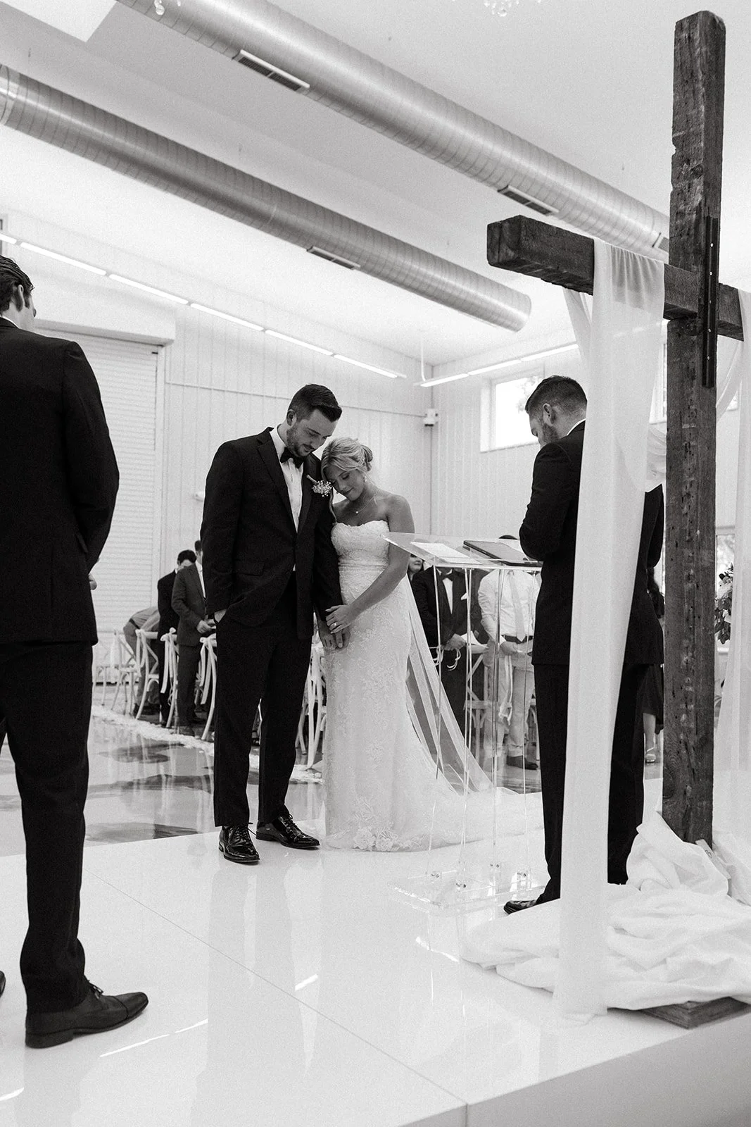 A black-and-white photo of a wedding ceremony with the bride and groom holding hands and bowing their heads, standing beneath a wooden cross with draped fabric, while a person on the right reads from a book and guests seated in the background look on