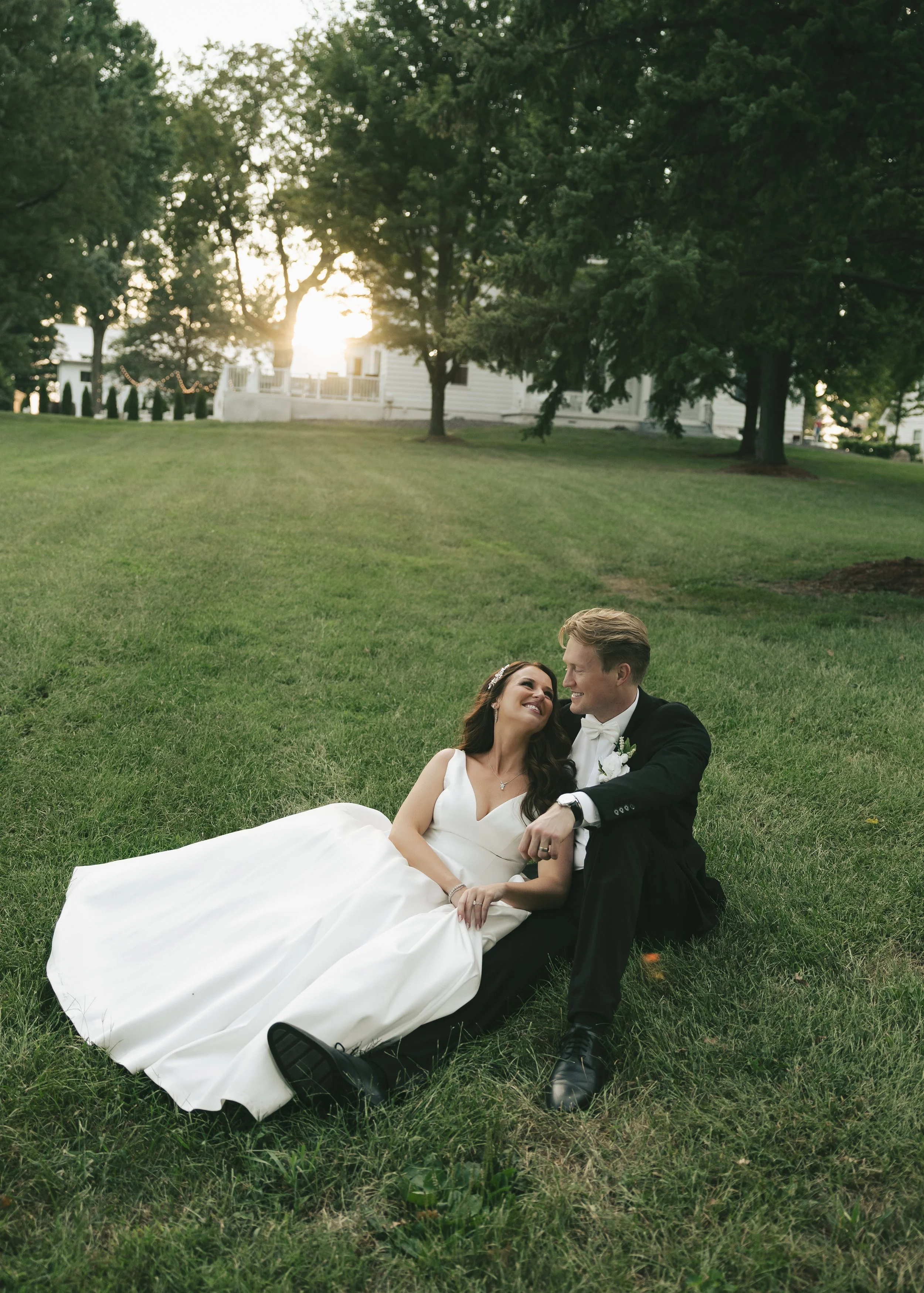 A bride and groom sitting on the grass in a park during sunset, smiling and looking at each other, with trees and a white house in the background.