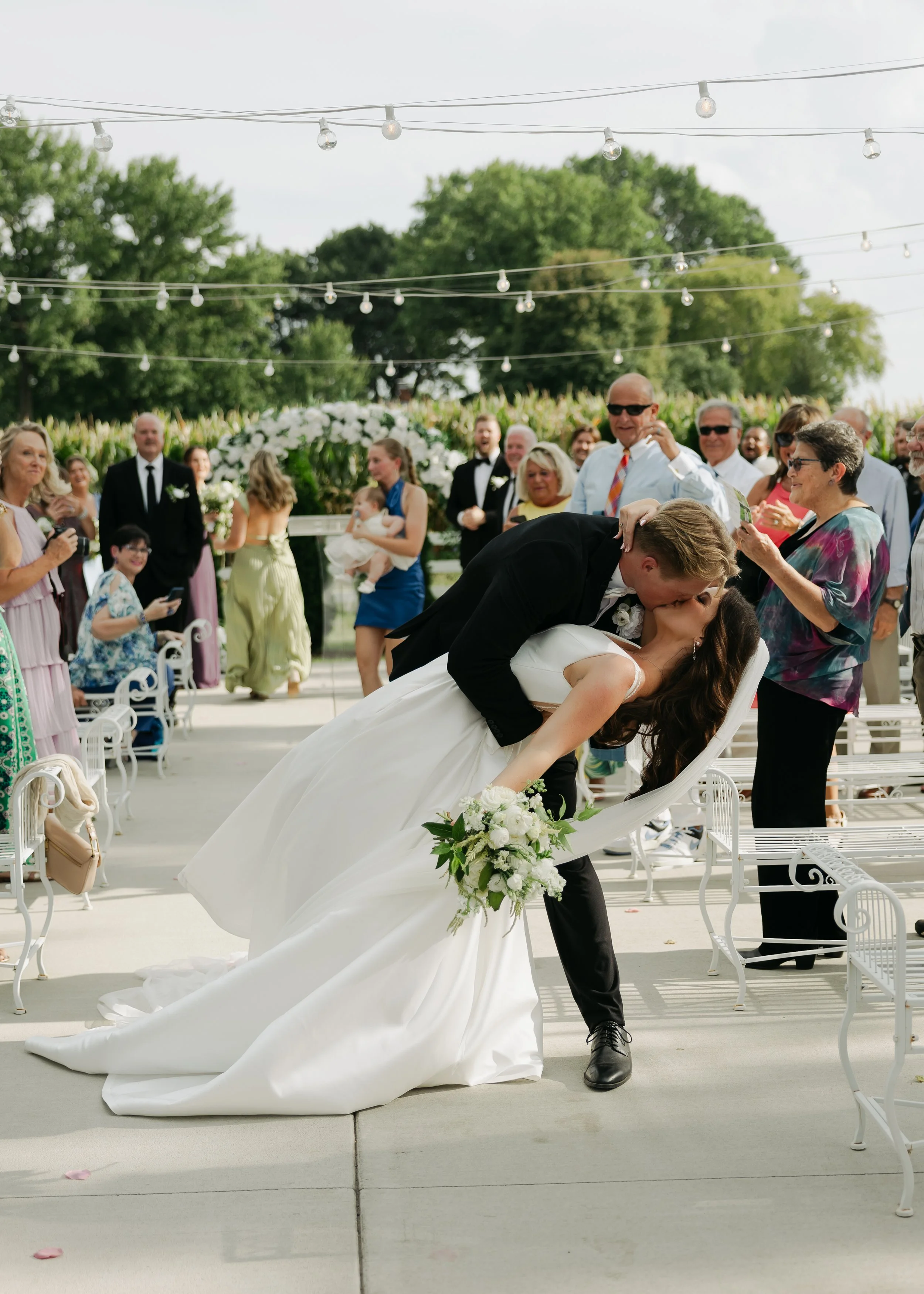 A newlywed couple kissing during their outdoor wedding ceremony, surrounded by guests, with string lights overhead and greenery in the background. Central Illinois wedding venue Pole Barn Chic. Good for Southern Illinois wedding venue, too. 