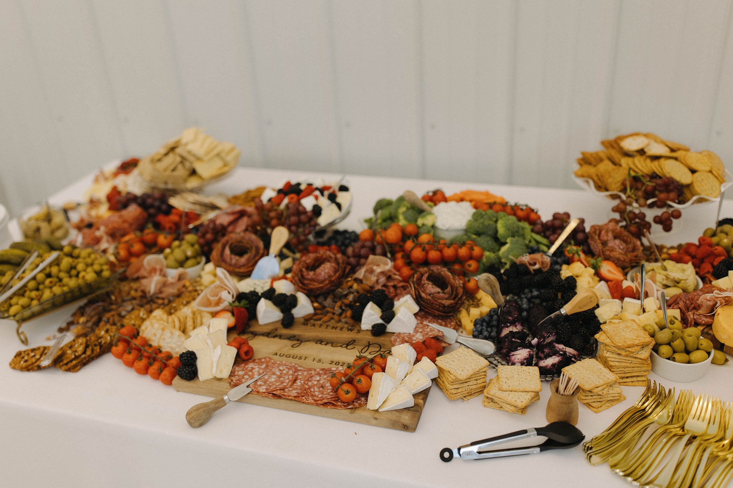 An assortment of cheeses, meats, fruits, vegetables, crackers, and snacks arranged on a table for a party or event, with a sign indicating a wedding date.