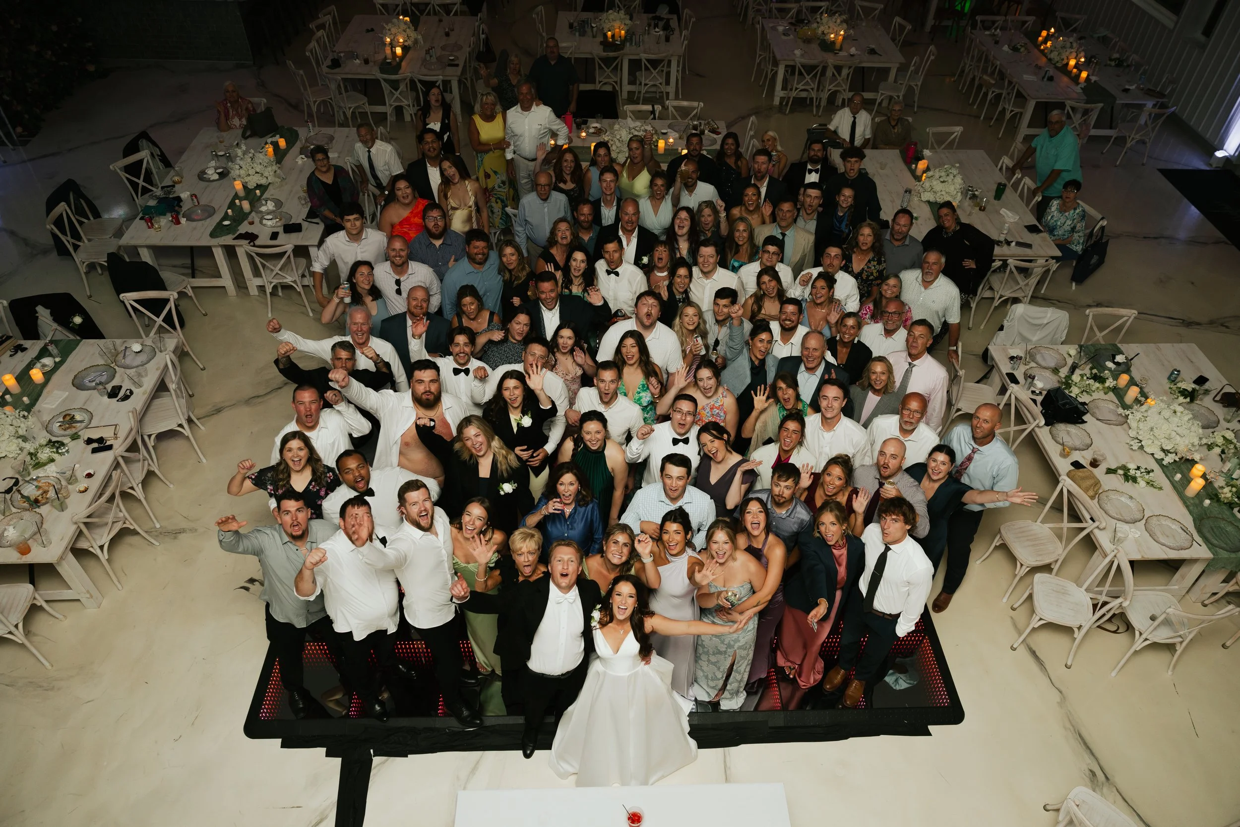 A large group of people at a formal event or celebration, gathered around tables decorated with candles and flowers, with some individuals smiling and celebrating.