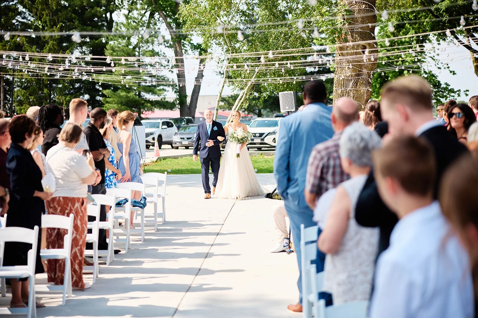 Bride walking down the aisle with her father at an outdoor wedding ceremony, surrounded by seated guests and decorated with string lights on trees.