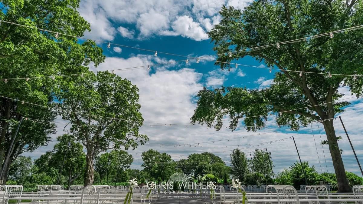 Outdoor event space with white chairs, string lights overhead, and large trees under a partly cloudy sky.