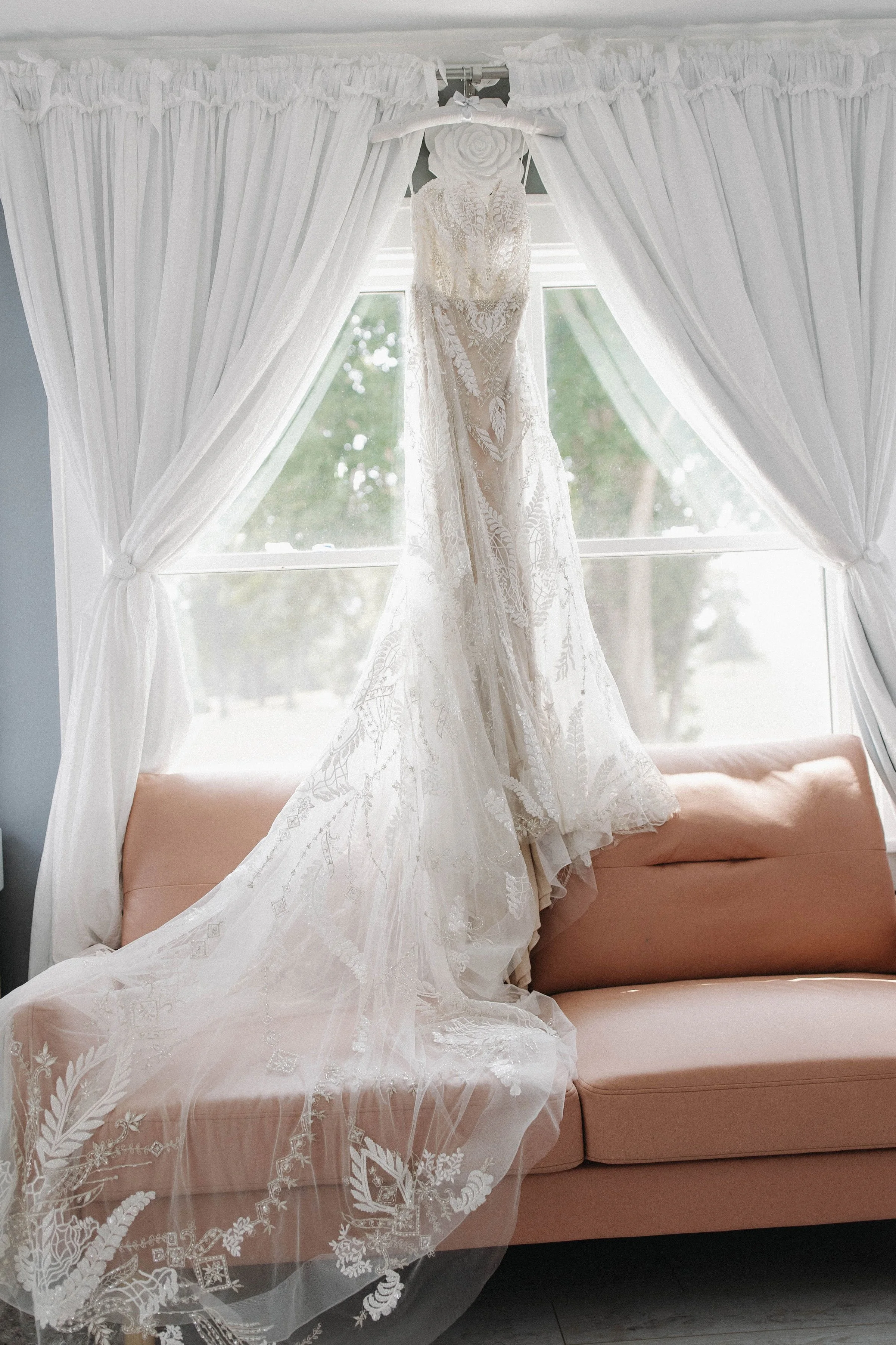 A wedding dress hanging in front of a window with white curtains, gold-colored sofa and a view of green trees outside.
