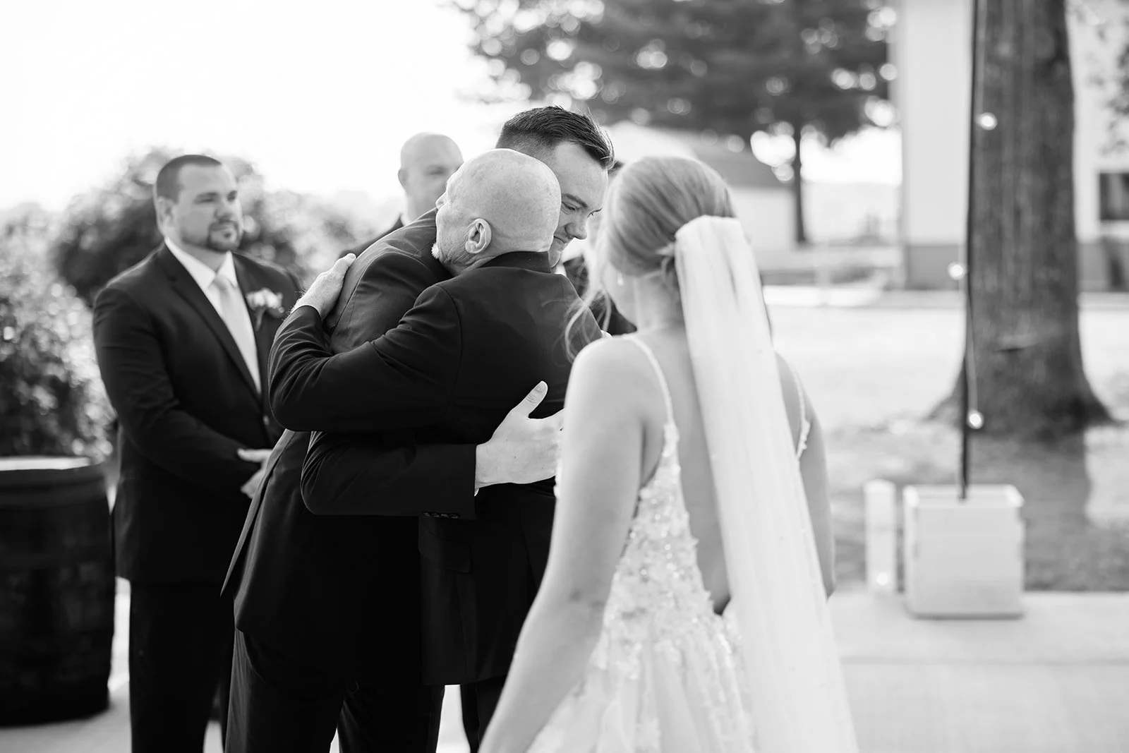 A black and white photograph of a wedding ceremony outdoors. The bride and groom are embracing each other, with the bride's back to the camera and the groom holding her. Two men in suits are standing behind them, witnessing the moment.