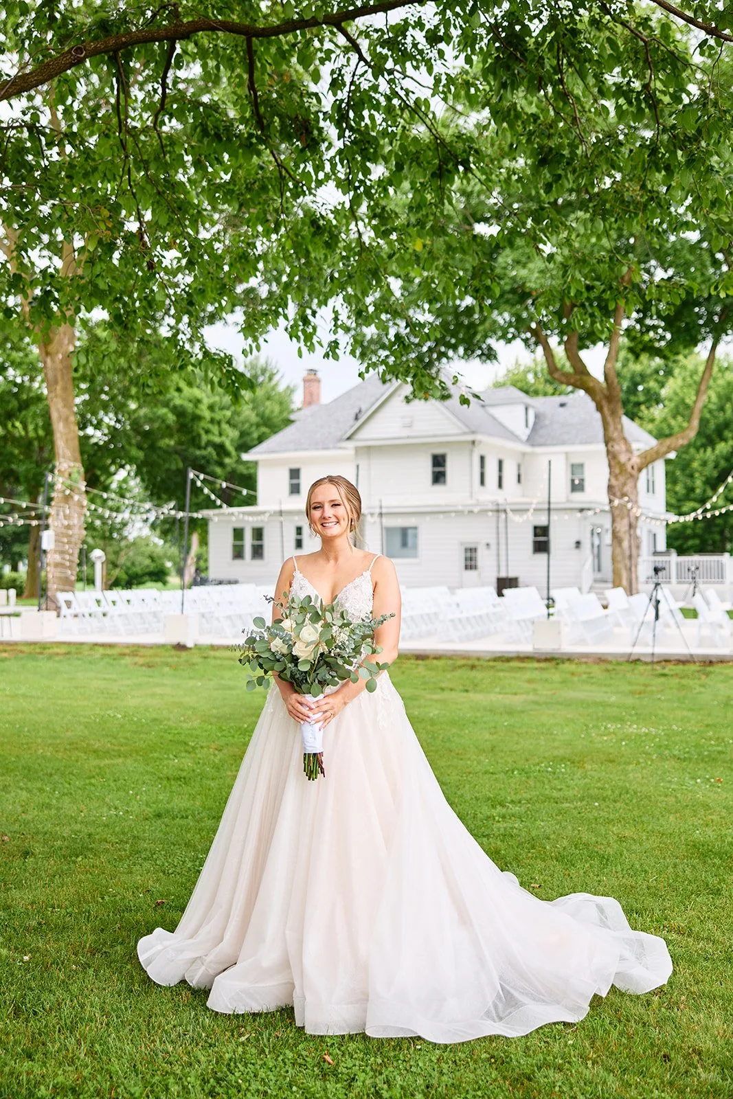 A bride in a white wedding gown holding a bouquet of white roses and greenery, standing outdoors on a lawn with trees and a large white house in the background, under string lights.