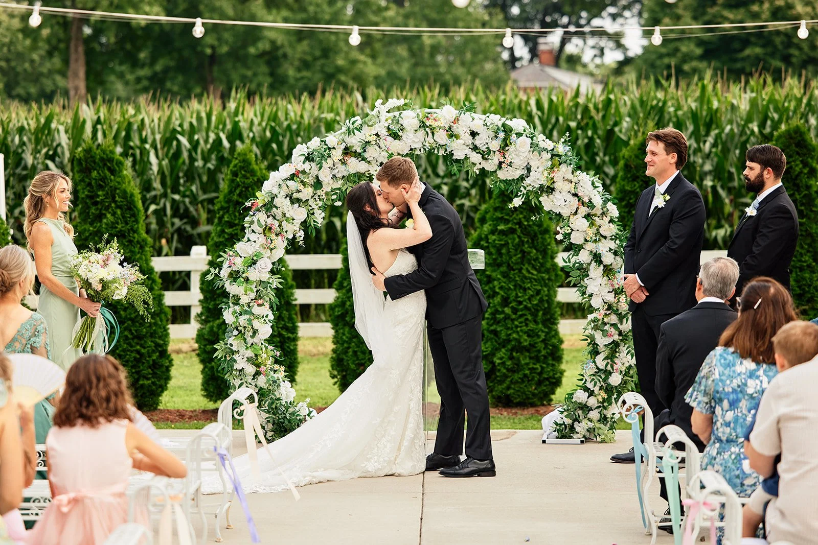 A bride and groom kiss under a floral arch during their outdoor wedding ceremony, with guests watching and a cornfield in the background.