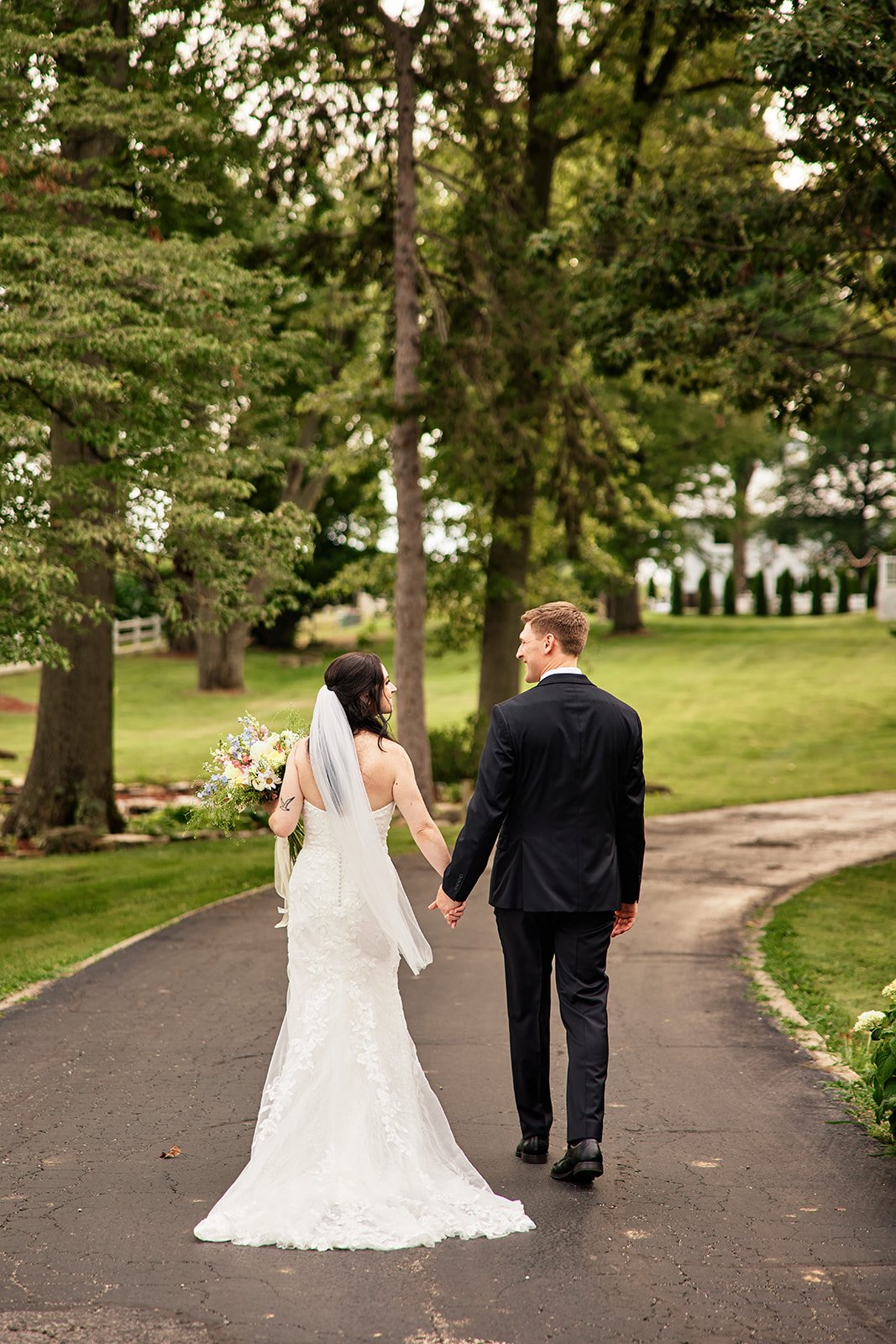A bride and groom walking hand in hand on a paved path in a green park, with trees and grass around them, photographed from behind.