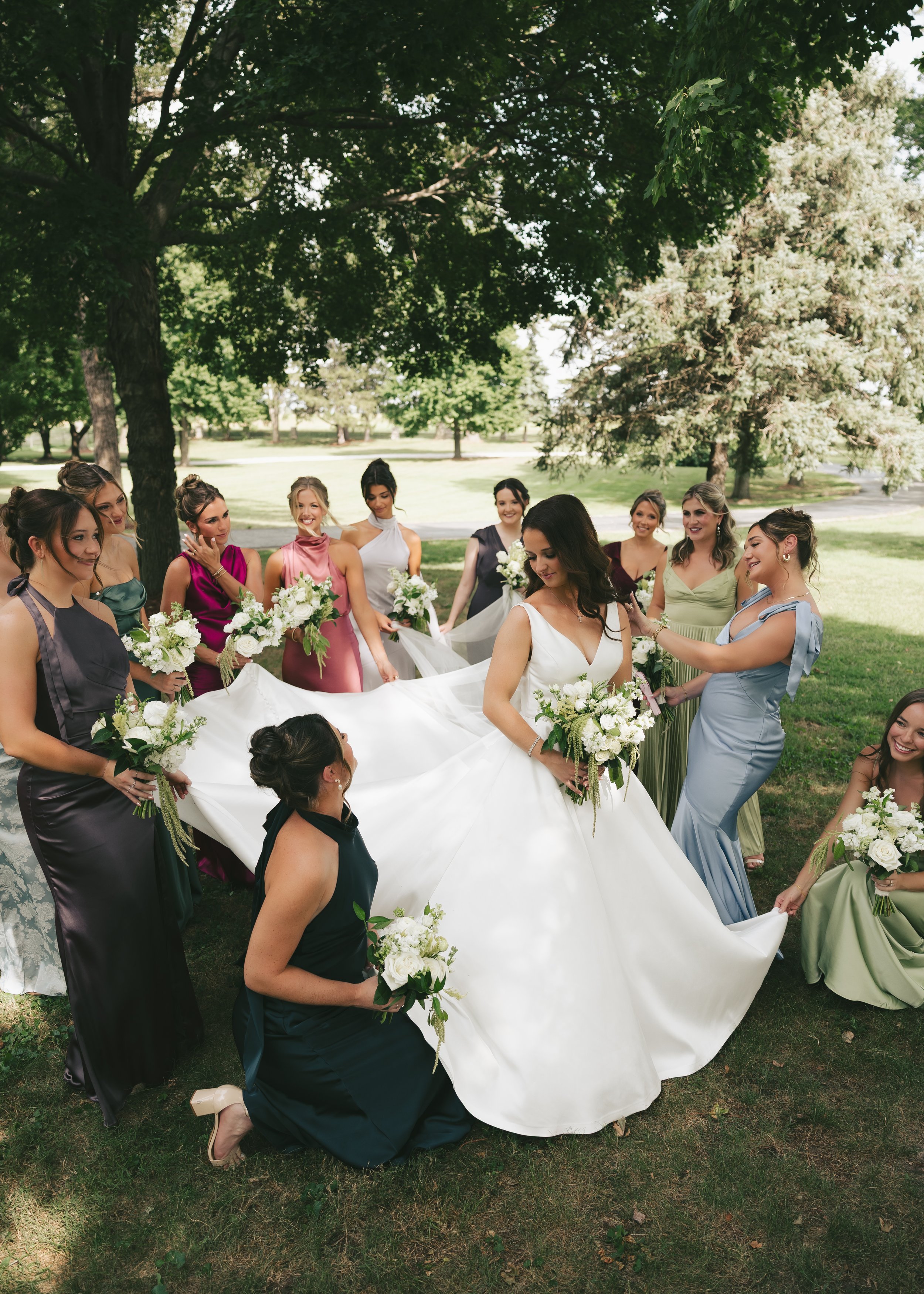 A group of women in colorful dresses gather outdoors under trees at Pole Barn Chic in Springfield IL, holding white flowers and a white fabric, celebrating at a wedding venue.