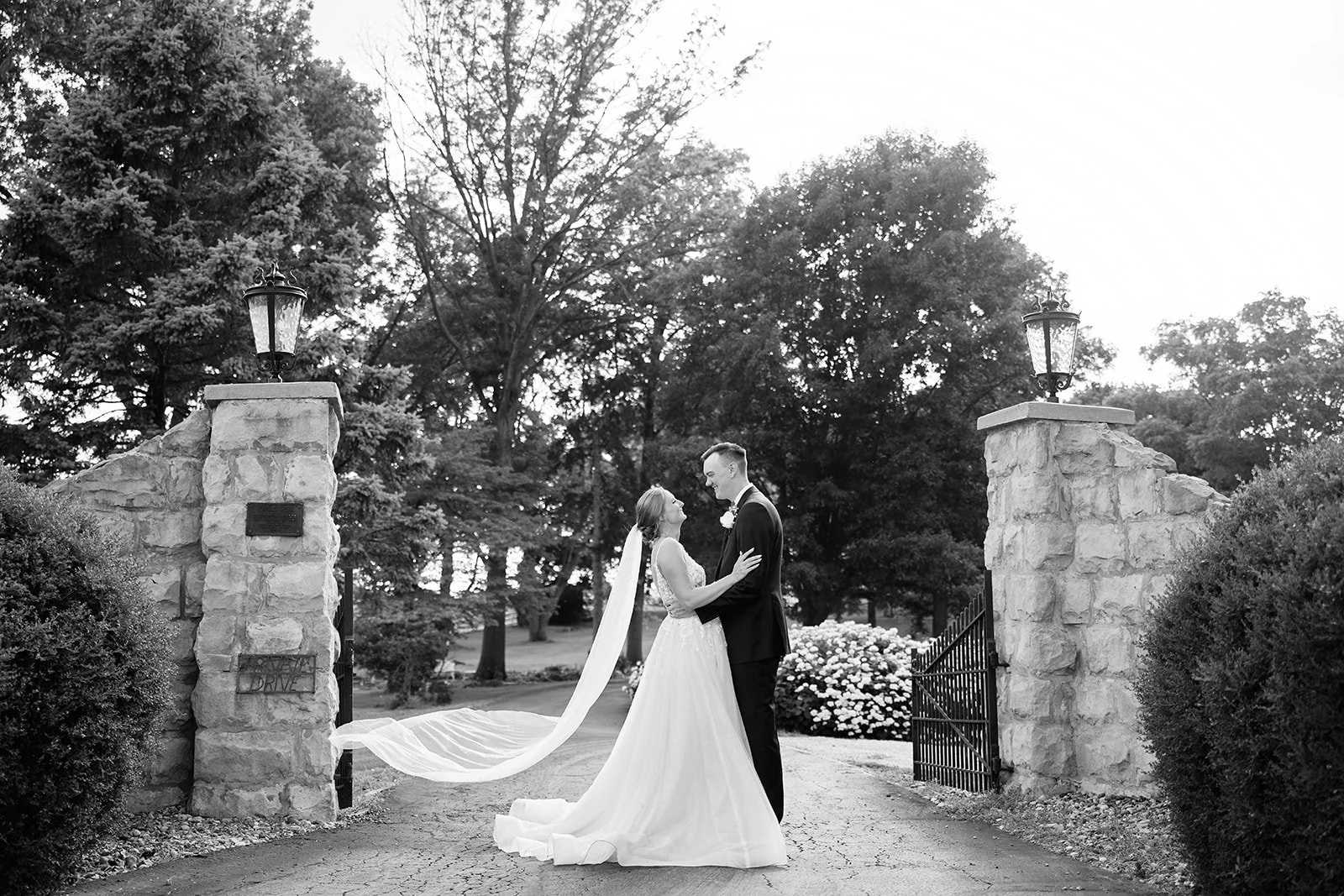 A couple in wedding attire, a bride in a white gown and veil and a groom in a tuxedo, standing and smiling at each other in an outdoor setting with trees and stone pillars and gates.