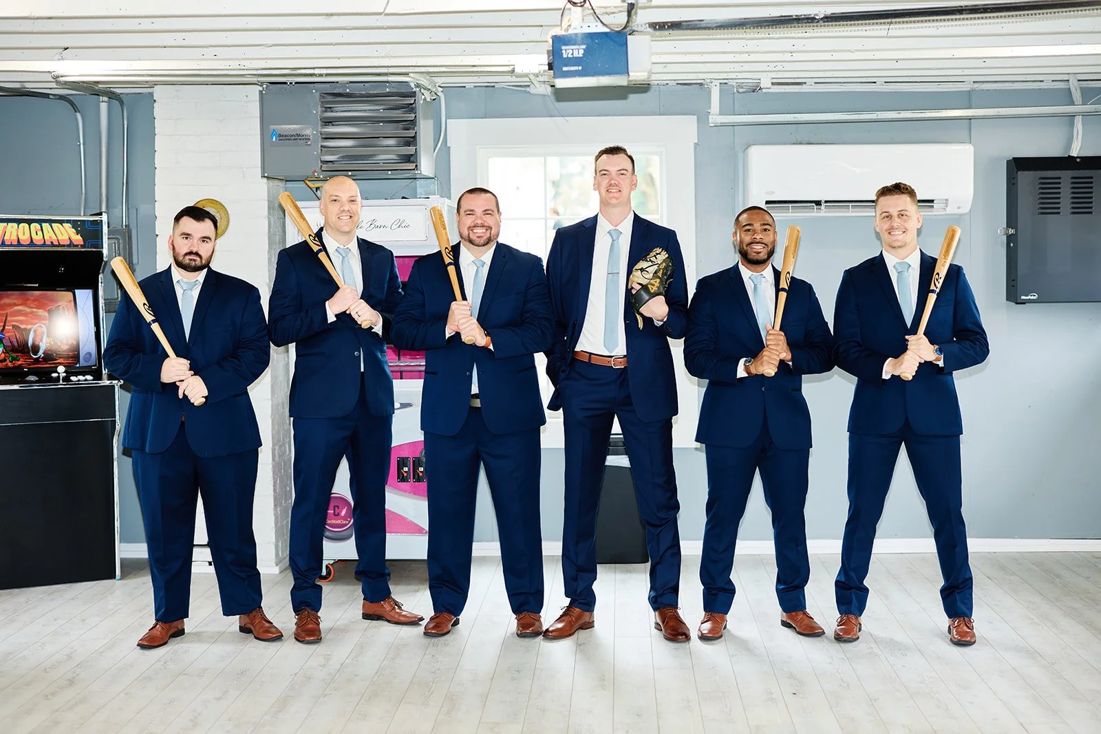 Six men in matching blue suits and light blue ties holding baseball bats, standing indoors with arcade games and air conditioning units behind them.