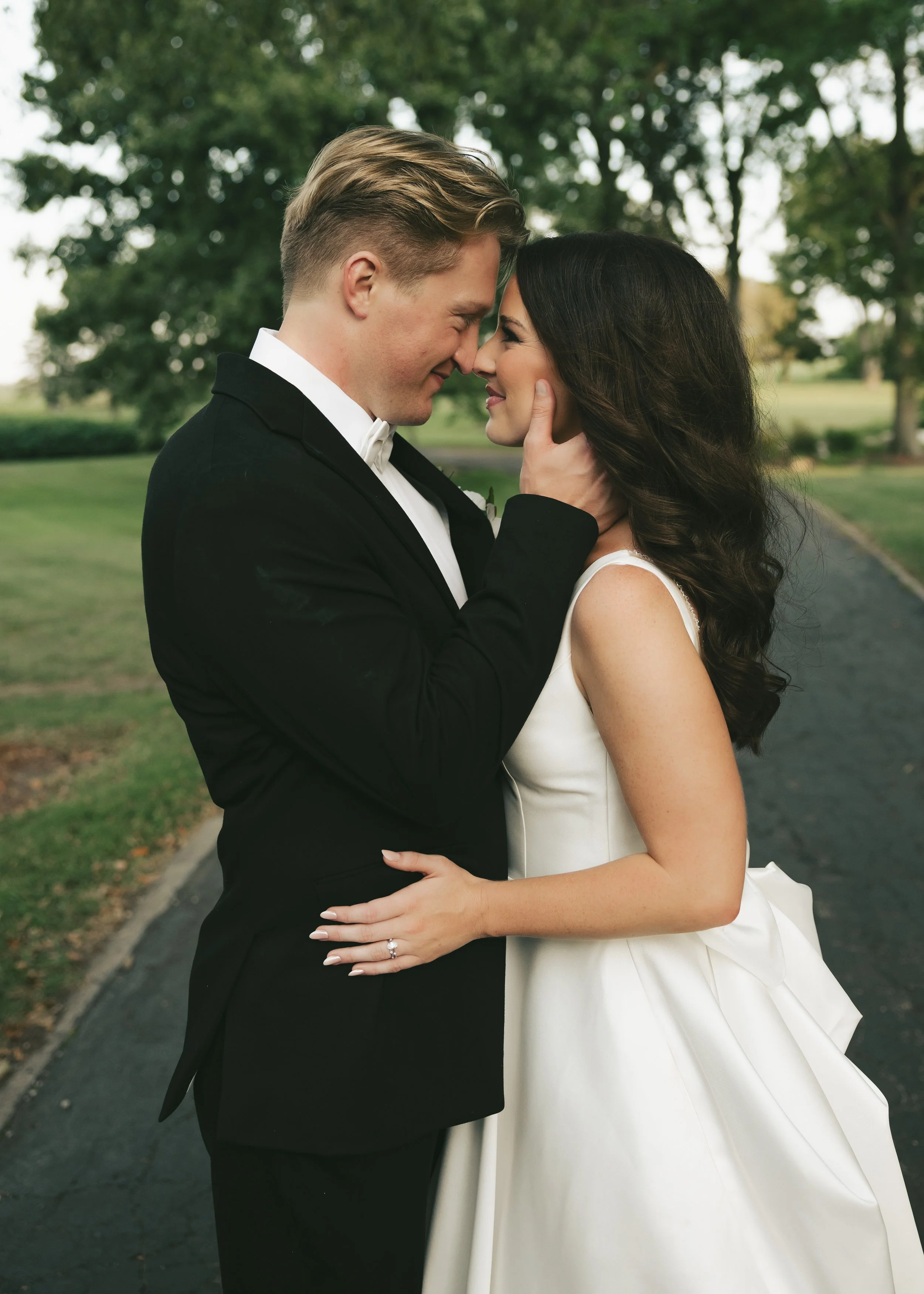 A bride and groom in wedding attire close together, touching foreheads and smiling, outdoors on a pathway with green trees in the background. Located at Pole Barn Chic in Springfield, IL. Central Illinois Southern Illinois