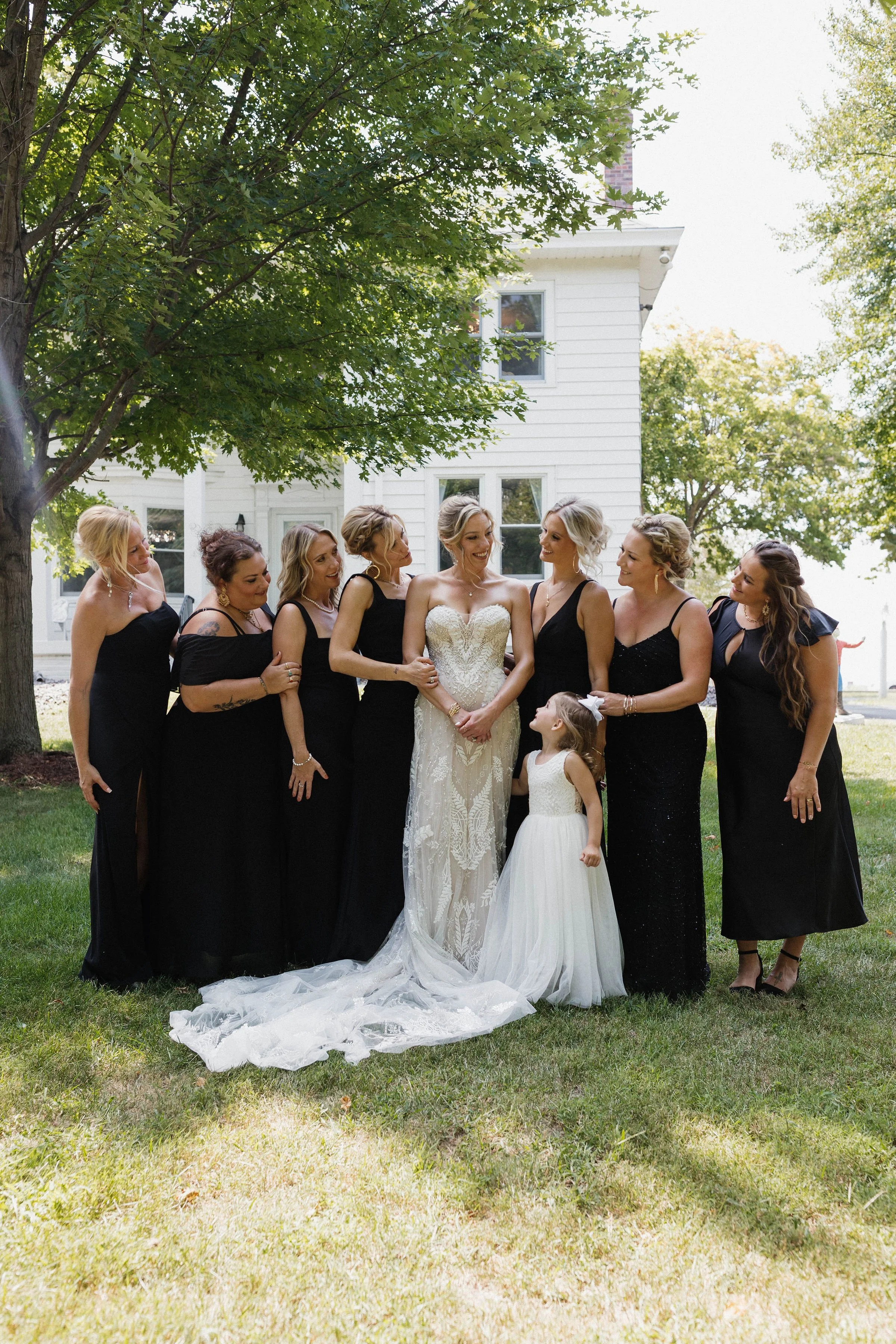 Bride in a white wedding dress surrounded by seven bridesmaids and a young girl in a white dress, all standing on grass near a white house with trees in the background.