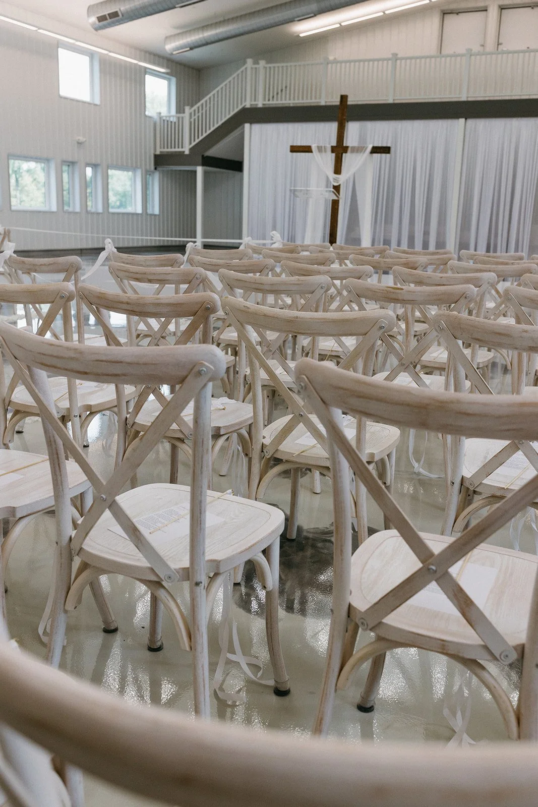 Empty chairs set up for a wedding ceremony with a cross and white curtains in a barn-like venue.