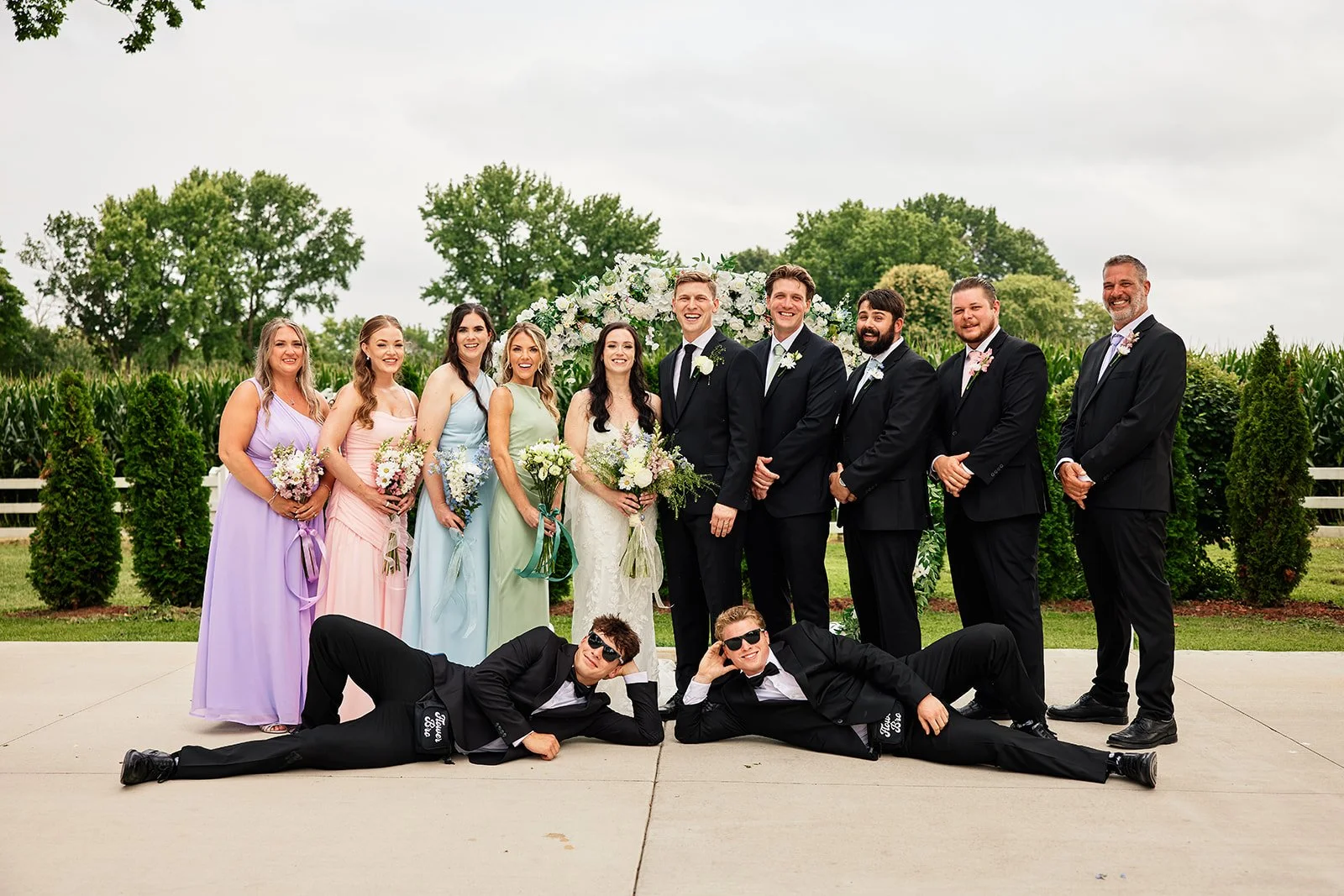 Group of wedding party members, including the bride and groom, bridesmaids, groomsmen, and two men in tuxedos, posing outdoors on a concrete area with greenery and trees in the background, during daytime. Flower Bros. Flower dudes. Bridal party.