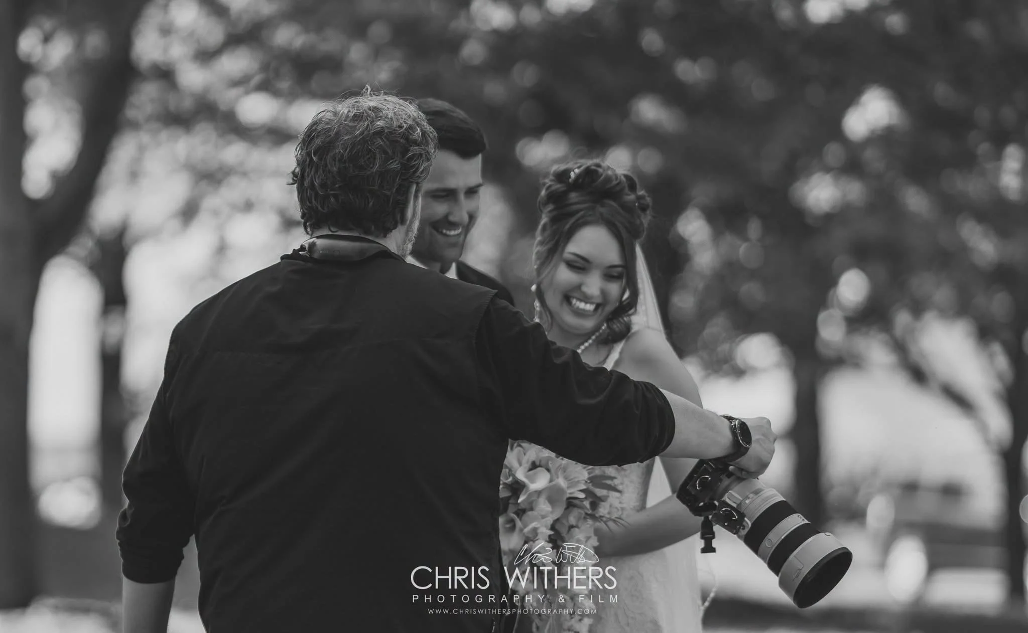 Three people outdoors, two men and a woman, smiling and looking at each other, with a camera in the woman's hand. Trees in the background, black and white photo.
