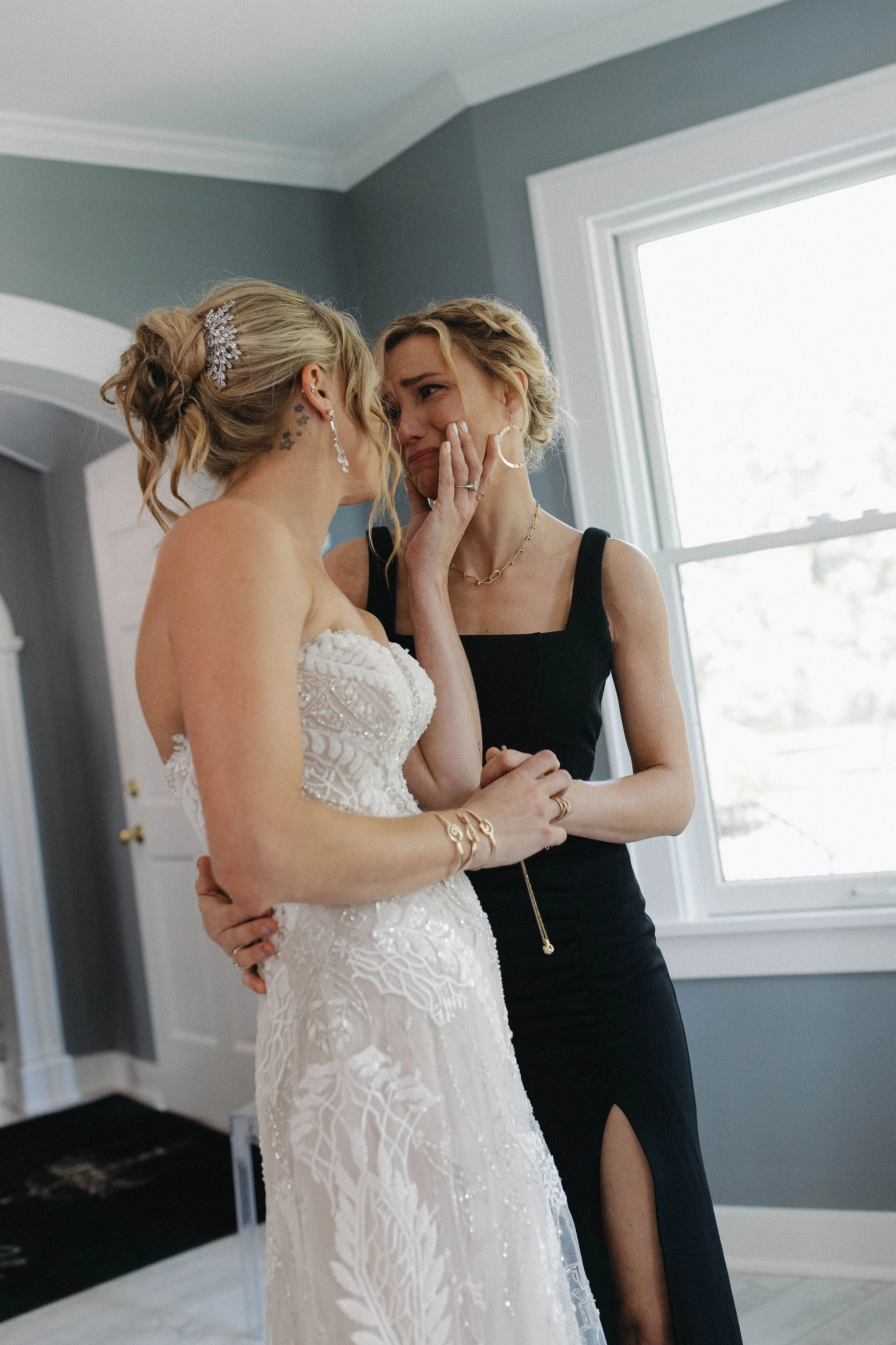 A bride and her bride'smaid share an emotional moment during a wedding, with the bride in a white lace wedding gown and the bridesmaid in a black dress, inside a bright room with a window.