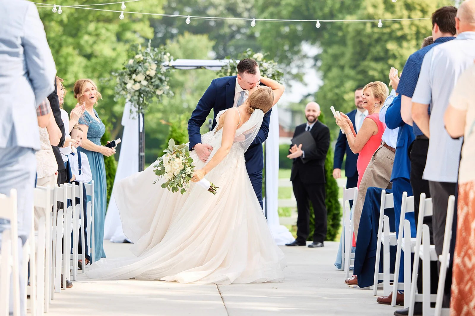 A newlywed couple sharing a kiss during their outdoor wedding ceremony, with guests cheering and clapping around them.