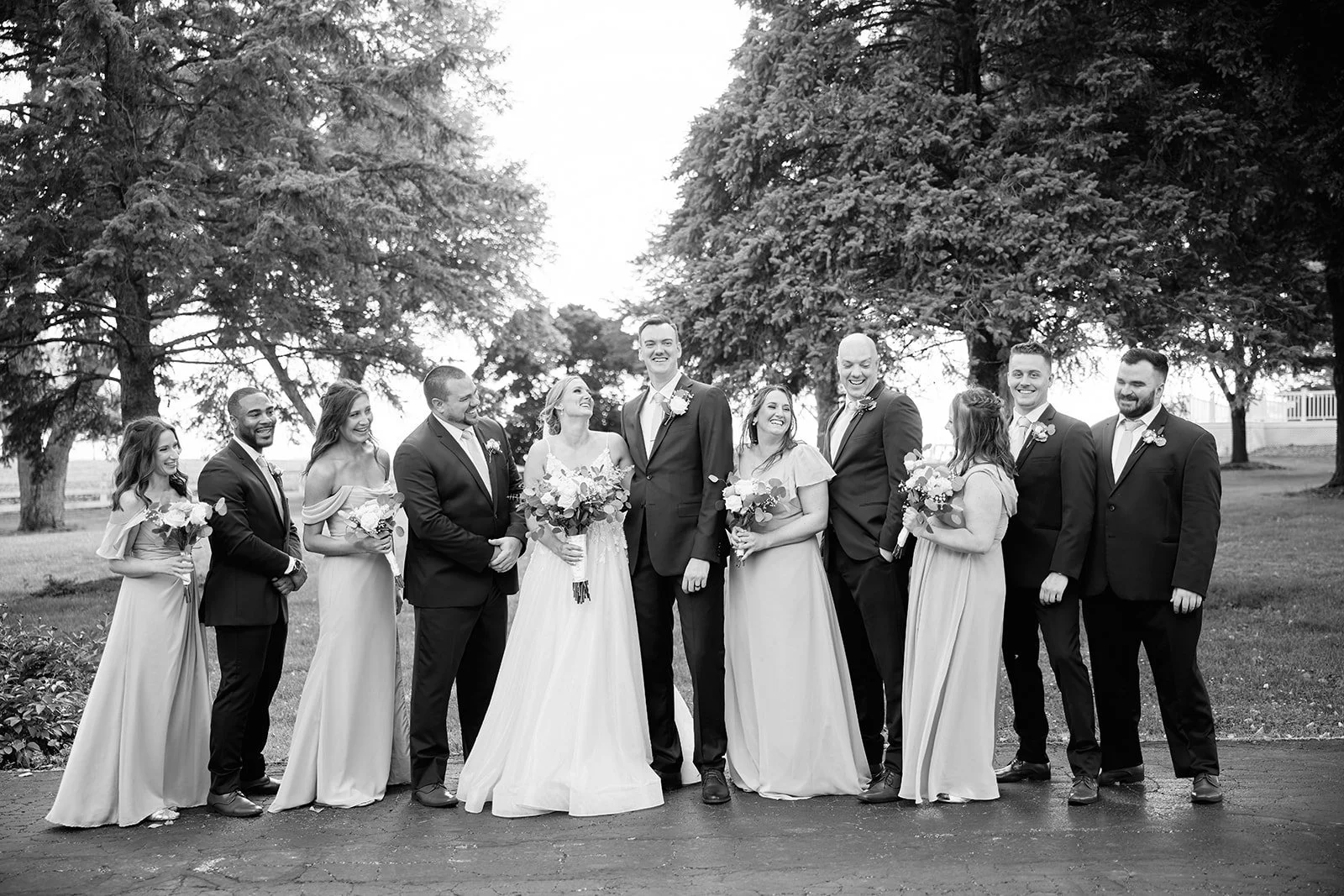 Black and white photo of a wedding party outdoors, with ten people, including the bride and groom, smiling and holding bouquets, surrounded by trees.