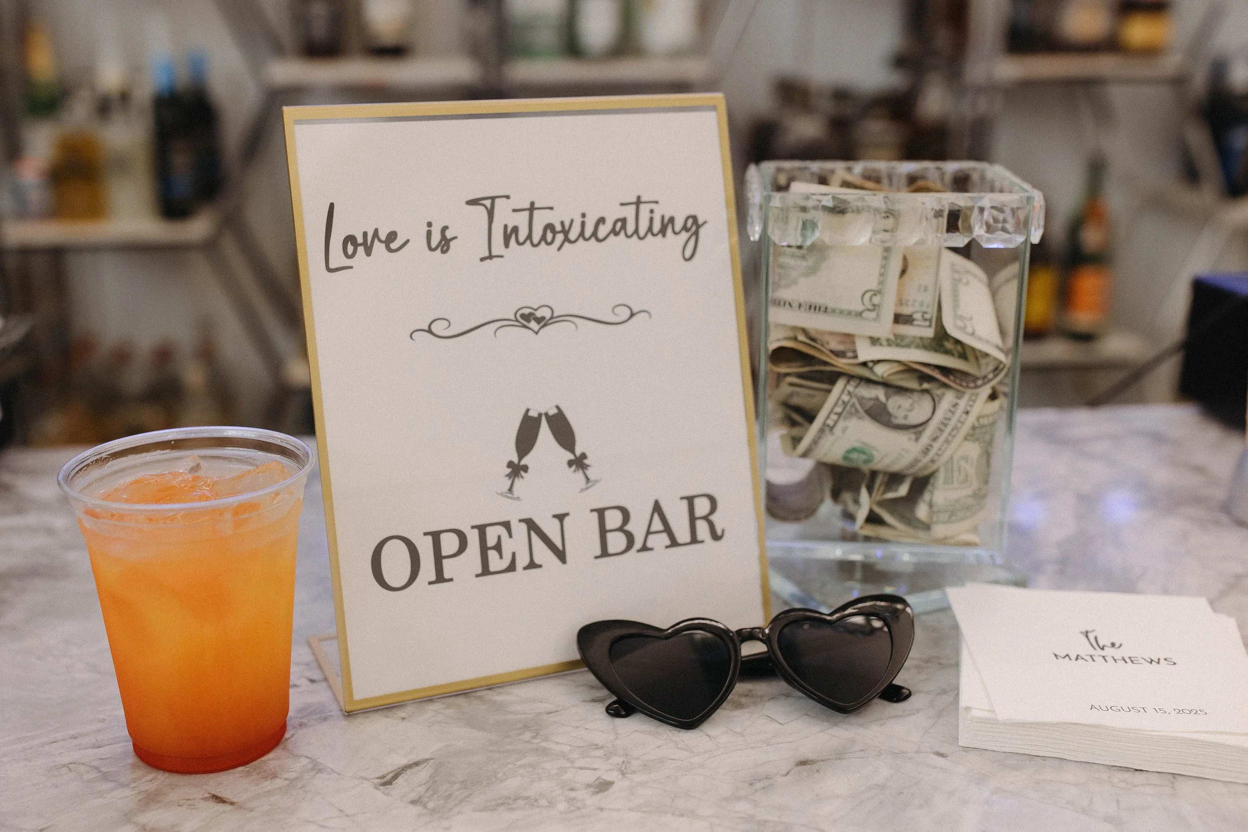 A drink with orange liquid and ice, a sign that reads 'Love is intoxicating, open bar,' a pair of heart-shaped sunglasses, a napkin with the words 'The Matthews' and date 'August 15, 2025,' a clear container filled with folded dollar bills.