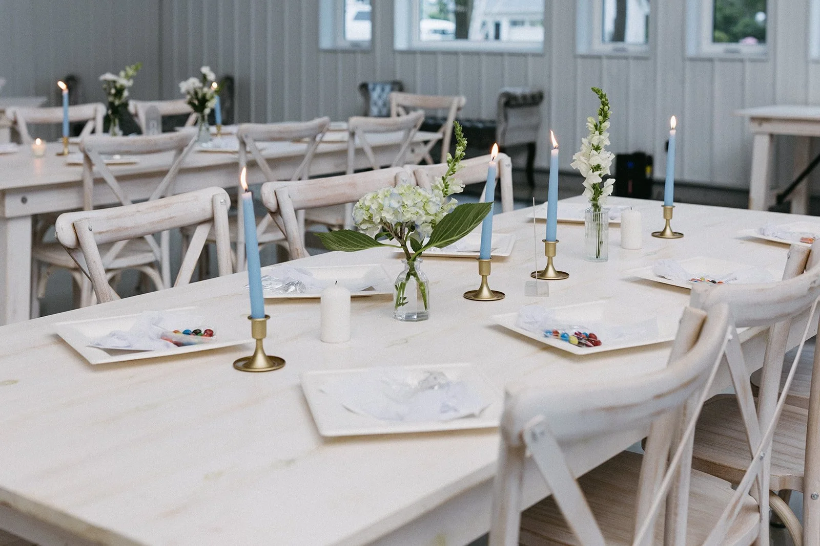 A decorated dining table set for a celebration with white plates, colorful candies, blue candles in gold holders, white flowers, and candles in white holders in a bright room with large windows.