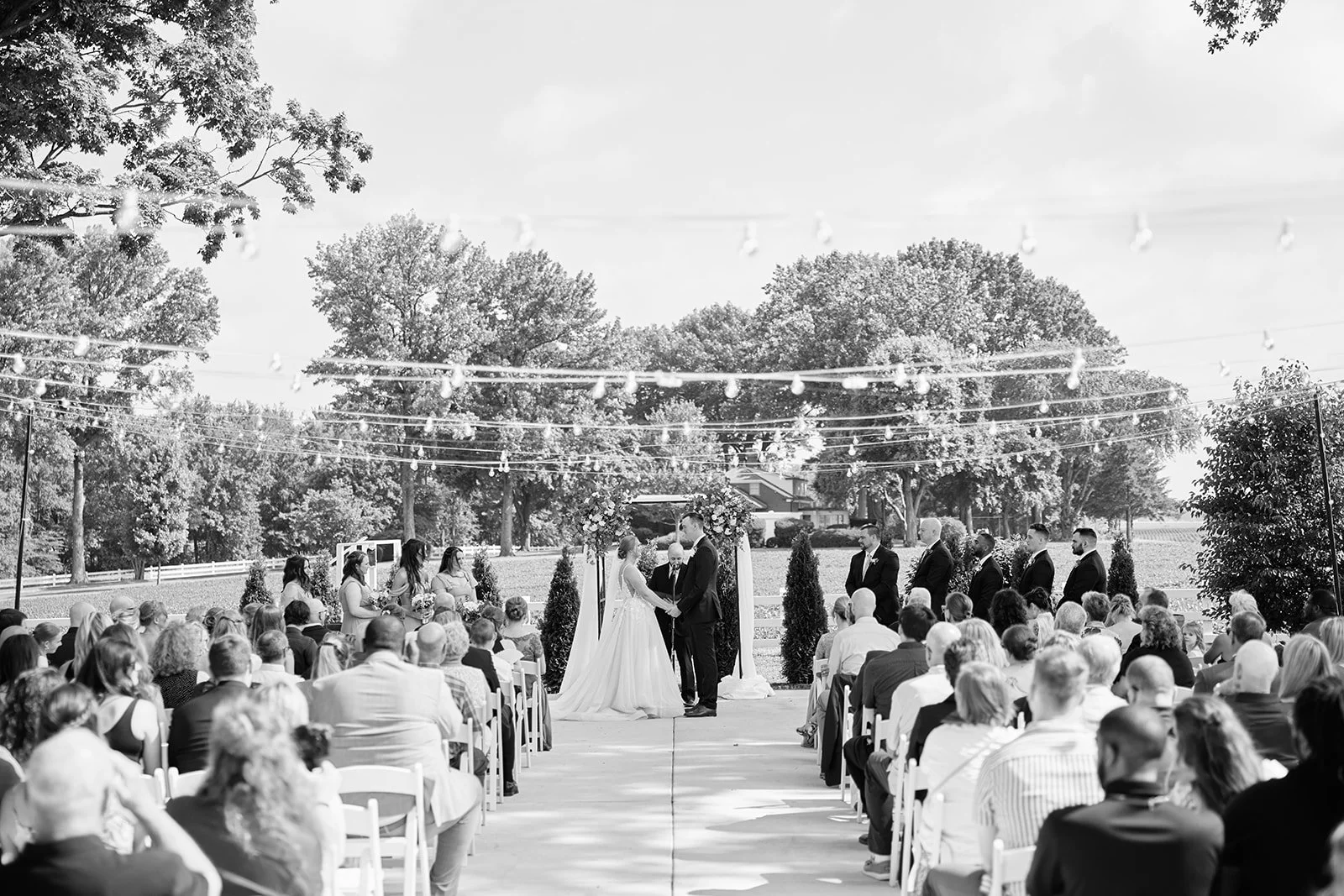 A black-and-white photo of a wedding ceremony taking place outdoors. The bride and groom are standing under a decorated arch, holding hands, surrounded by bridesmaids and groomsmen. Guests are seated in rows facing the couple, with trees and open sky