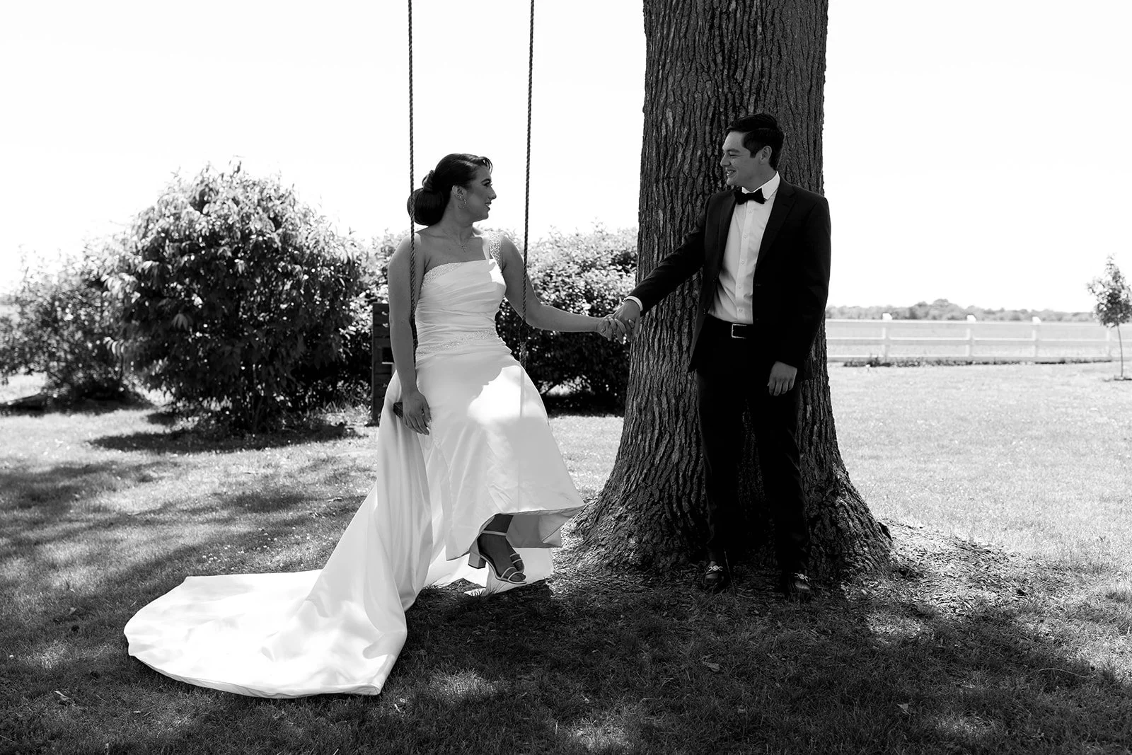 A black and white photo of the bride and groom holding hands at the tree, while the bride is in a swing.
