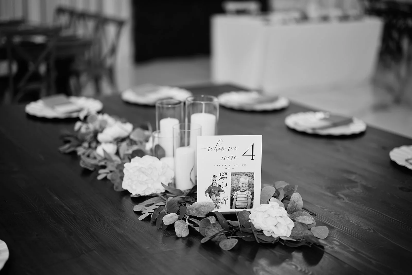 Black and white photo of a table decorated for a wedding, with candles, flowers, and a table number card displaying 'when we were 4' with photos of young children.