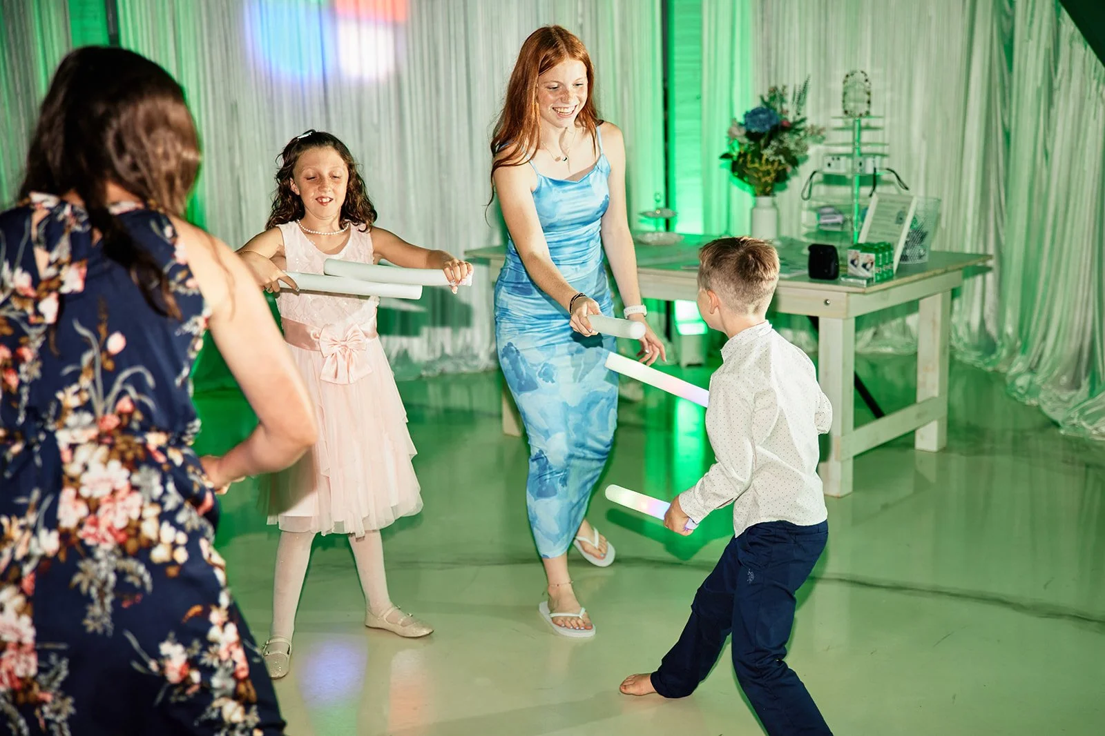 Women and children dancing with glow sticks at a party or celebration in a decorated room.