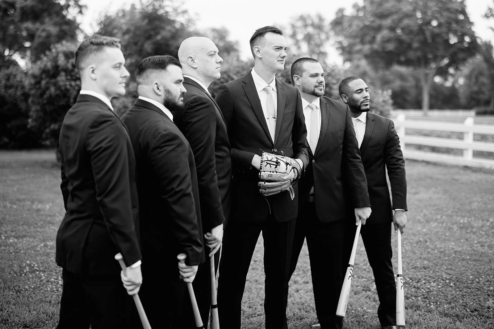 A group of six men in formal suits standing outdoors, some holding baseball bats, with one man in the middle wearing a baseball glove, suggesting a baseball team pose.
