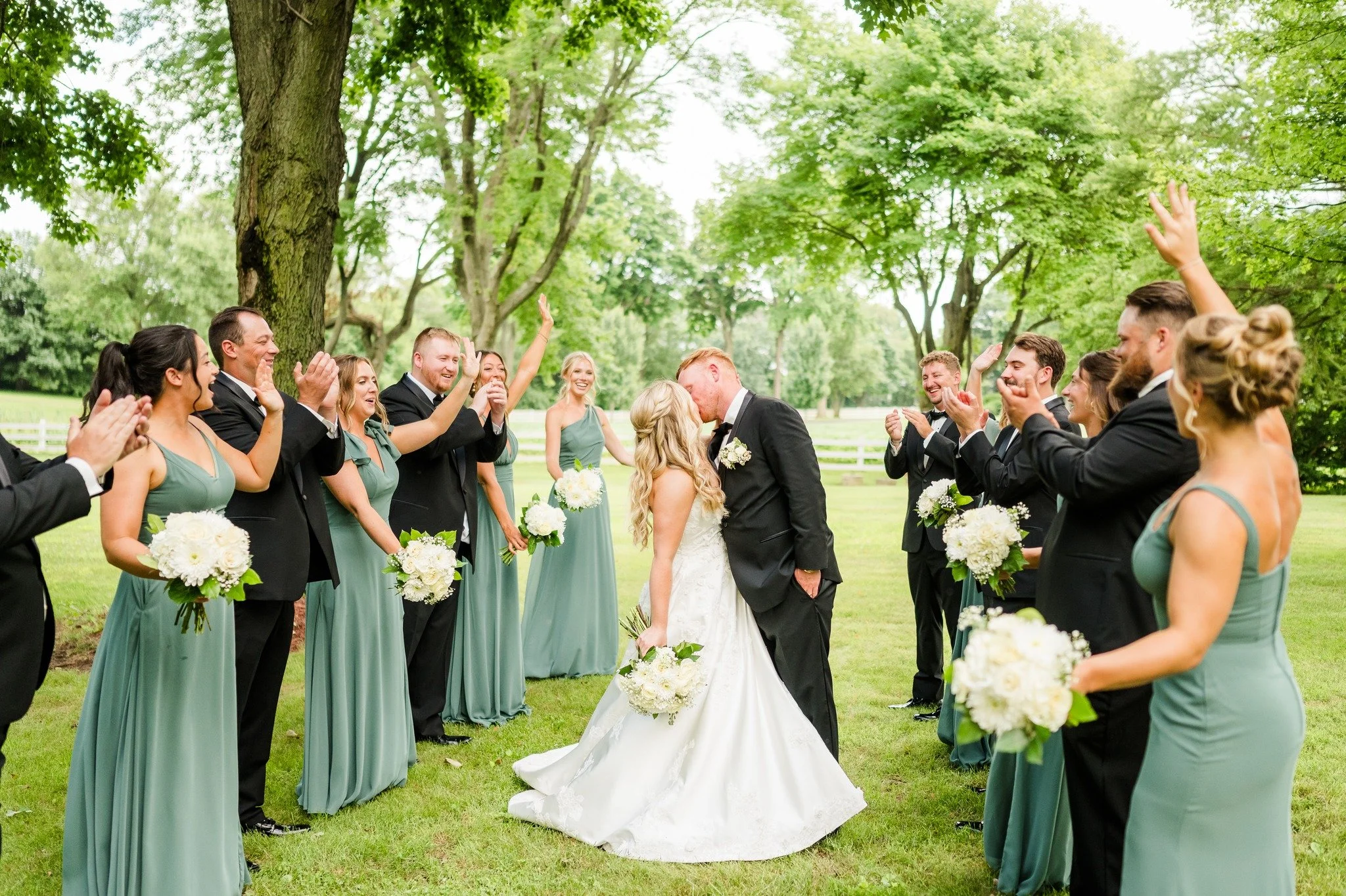 Couple kissing at an outdoor wedding ceremony surrounded by wedding party members holding bouquets, standing on grass with trees in the background.
